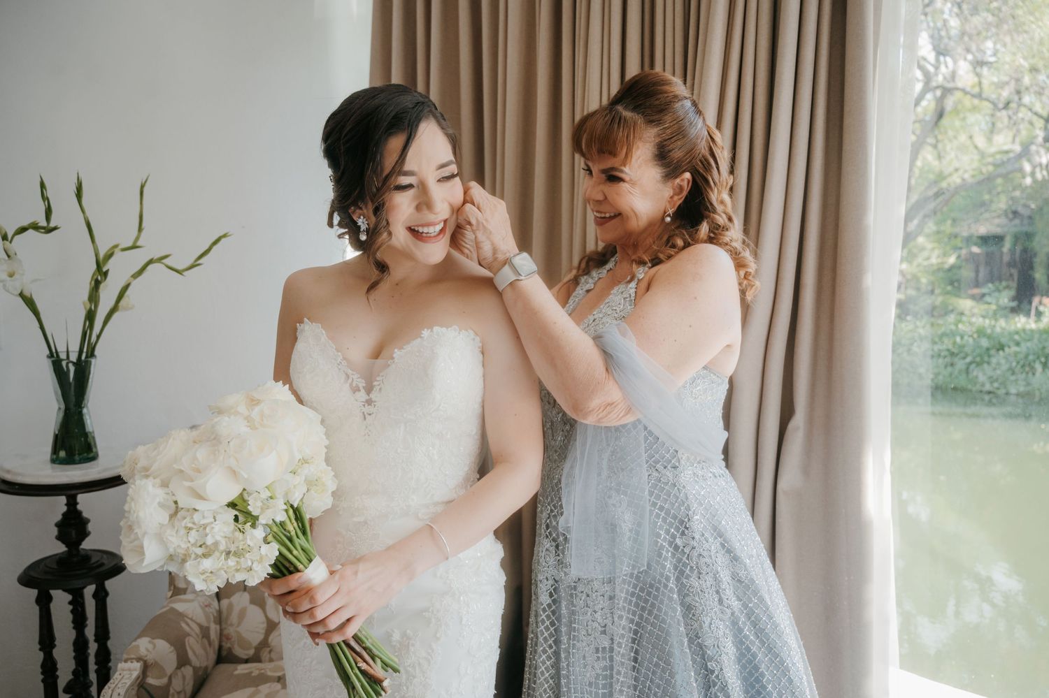 Bride in white strapless gown holds bouquet while woman in blue lace dress adjusts her earring near window.