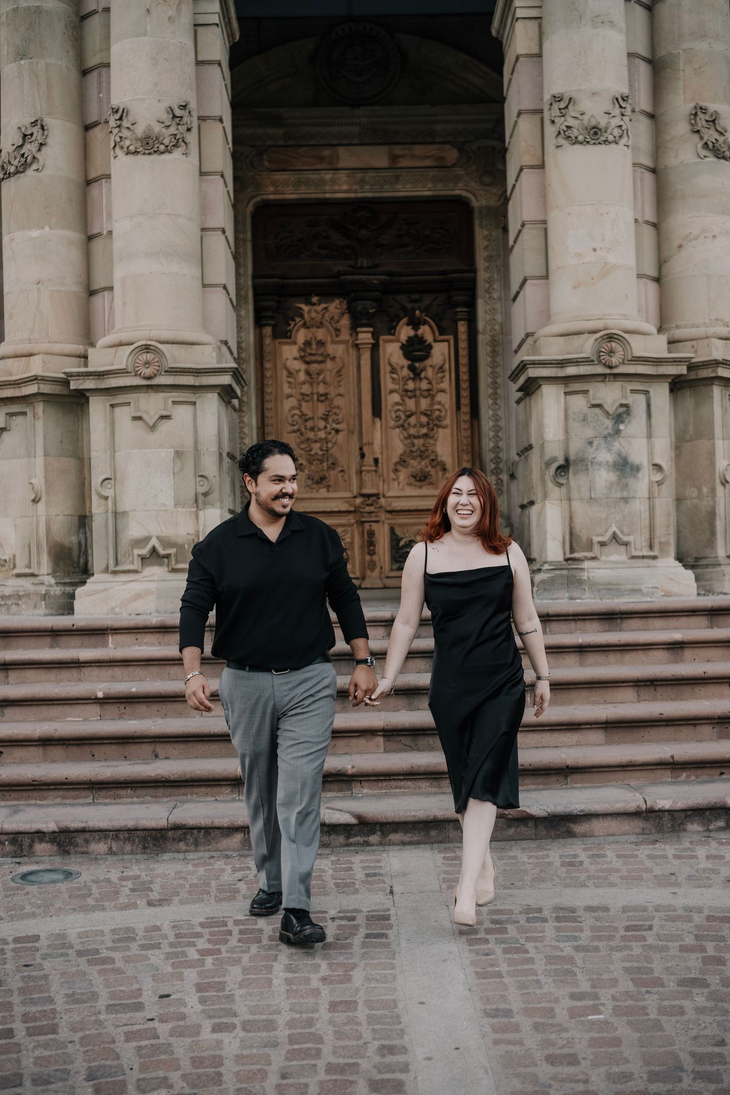 A couple holding hands and smiling while walking in front of an ornate historic building with carved wooden doors.