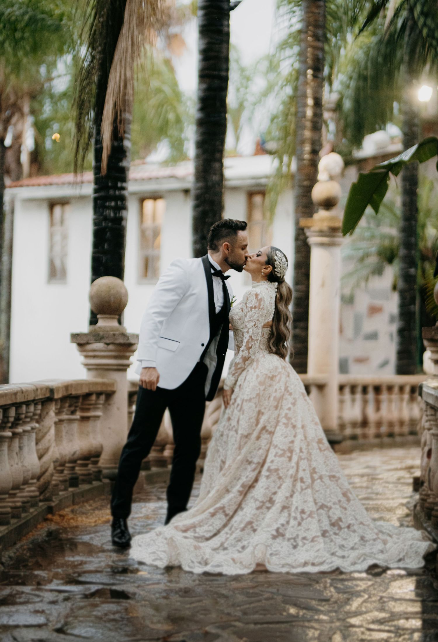 Bride and groom kissing on rainy terrace with palm trees and elegant architecture.