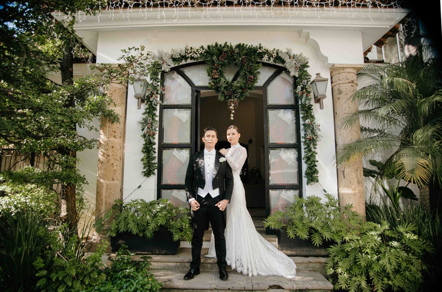 Elegant couple posing in wedding attire before a vine-covered villa entrance surrounded by lush tropical greenery.