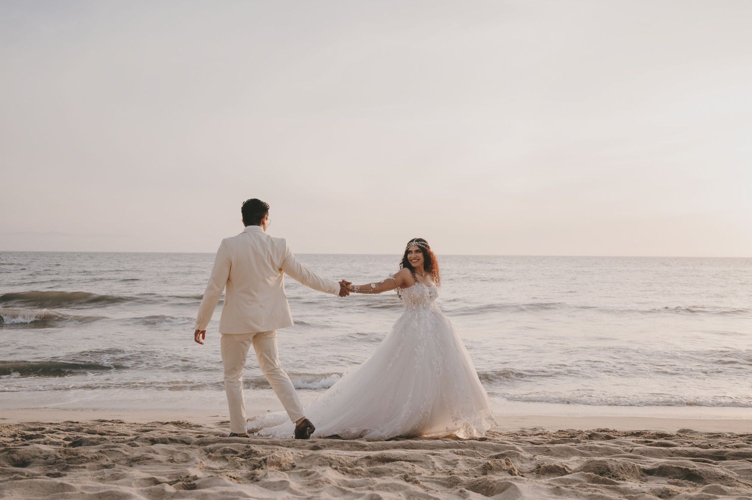 Bride and groom holding hands dancing on a sandy beach at sunset in elegant white wedding attire.