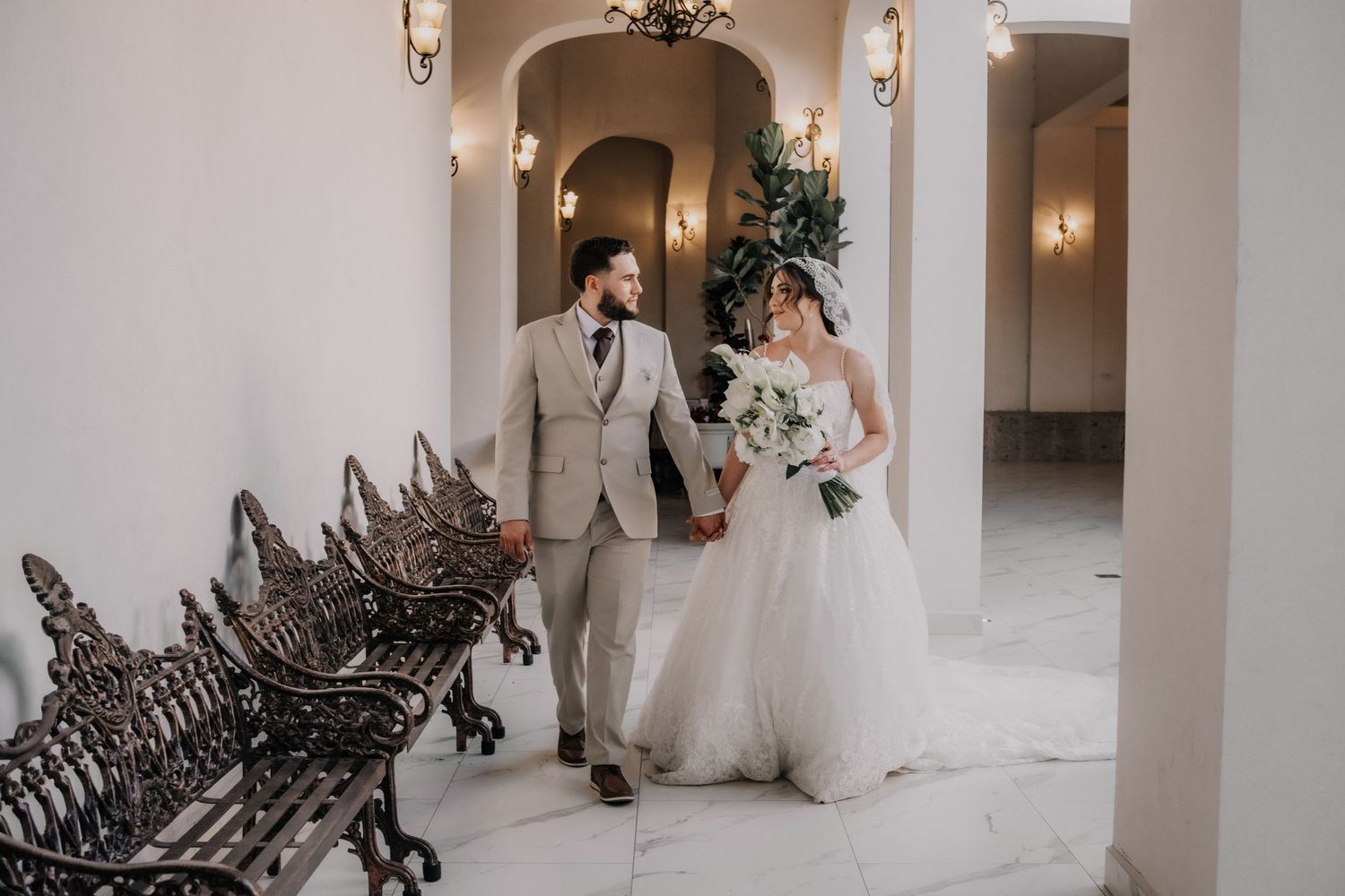 Bride and groom walk hand-in-hand through an elegant white hallway with ornate iron benches and warm wall sconces.