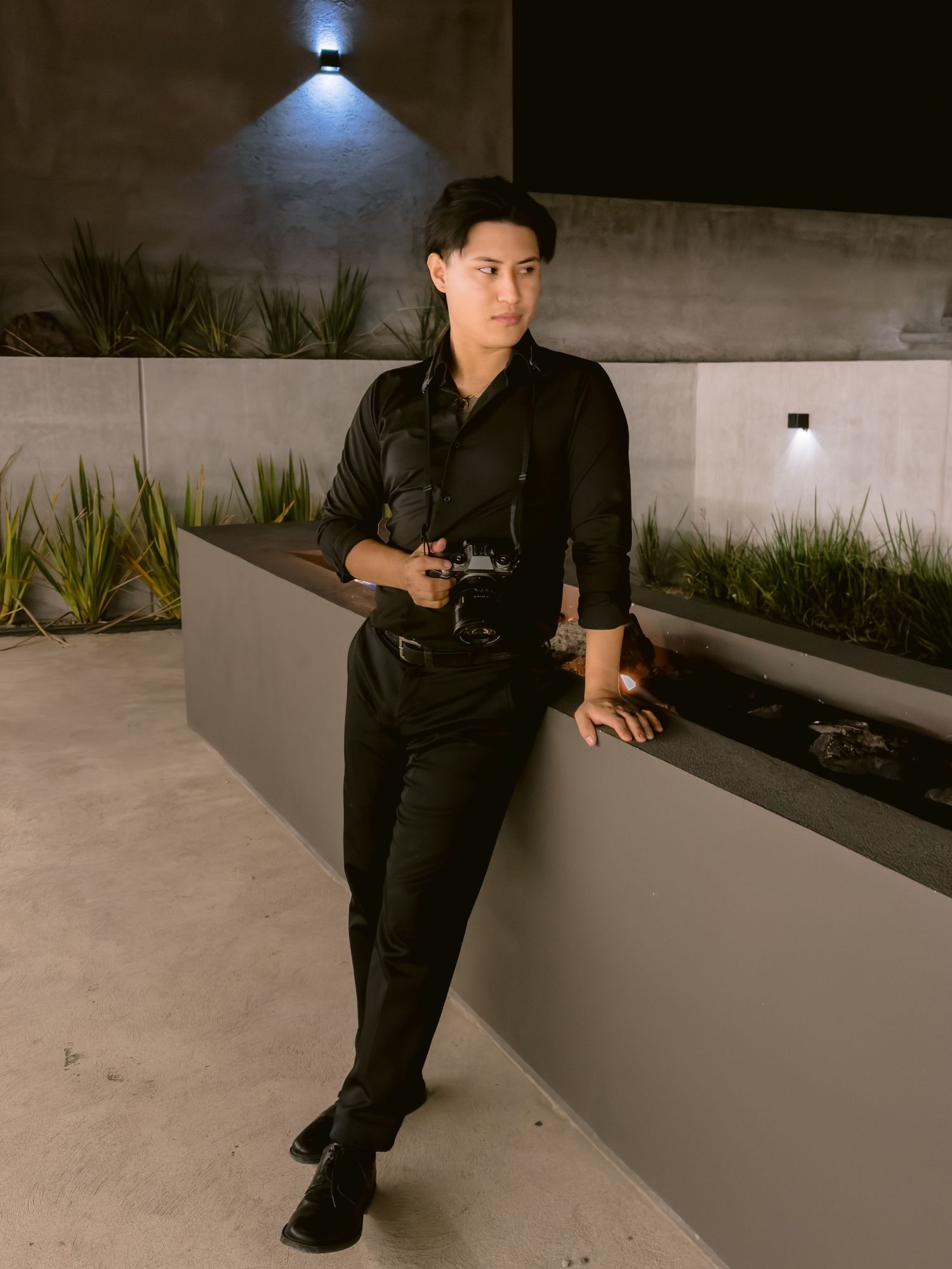 Young man in all-black outfit leaning against concrete wall at night, holding a camera outdoors.