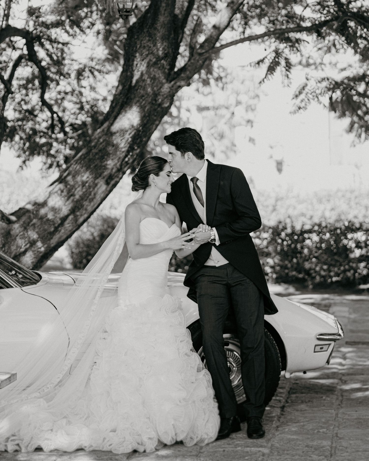 Black and white photo of bride and groom leaning against vintage car under trees.