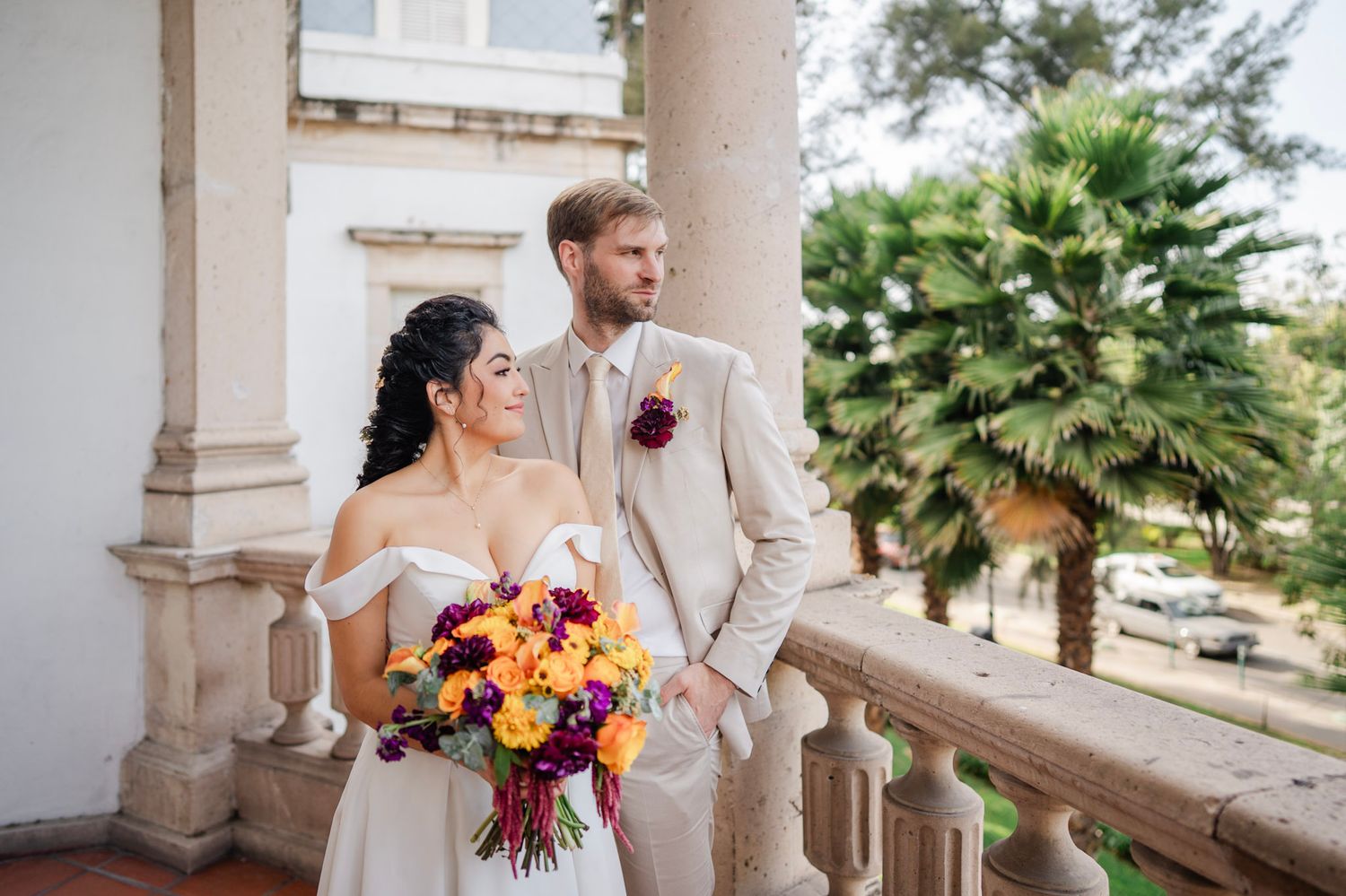 Bride and groom pose on a balcony with vibrant purple and orange bouquet, palm trees in background.