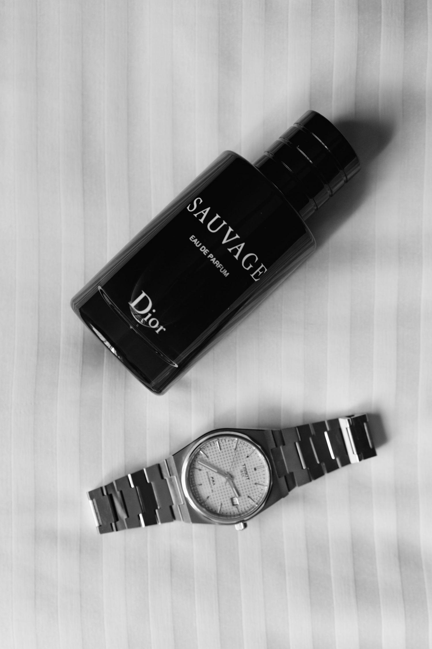 Black and white flat lay of Dior Sauvage Eau de Parfum bottle beside a silver luxury wristwatch on striped fabric.