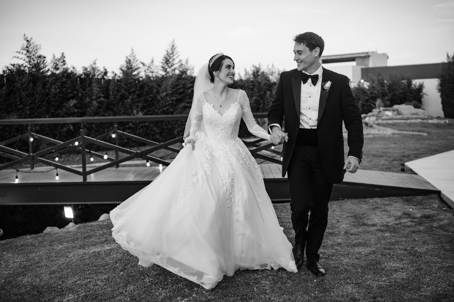 Black and white photo of a bride in a ball gown and groom in a tuxedo holding hands and walking outdoors at their wedding.