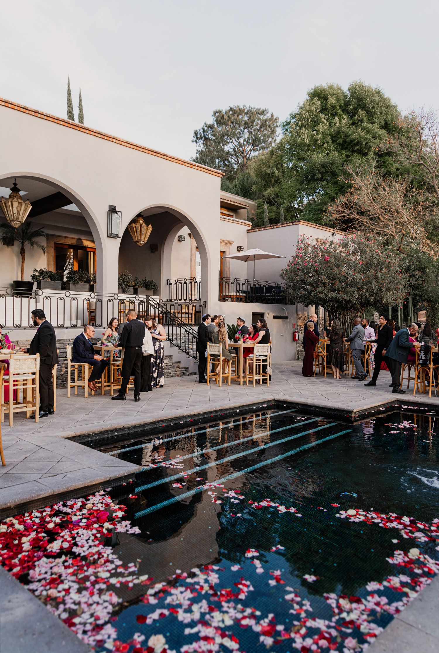Elegant outdoor wedding cocktail hour beside a rose petal-filled pool at a Spanish-style hacienda venue.