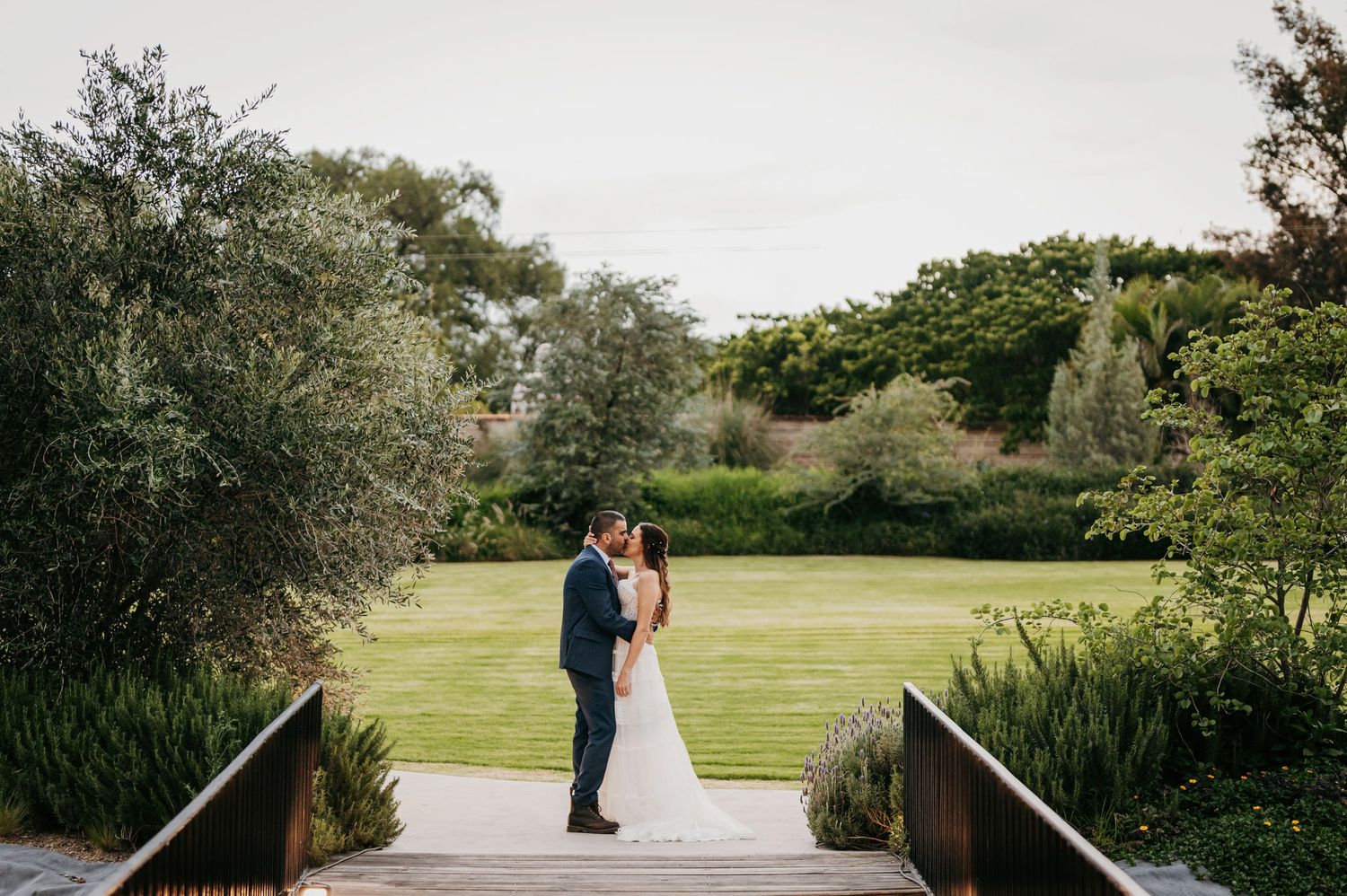Bride and groom share a romantic kiss on a wooden bridge surrounded by lush garden greenery.