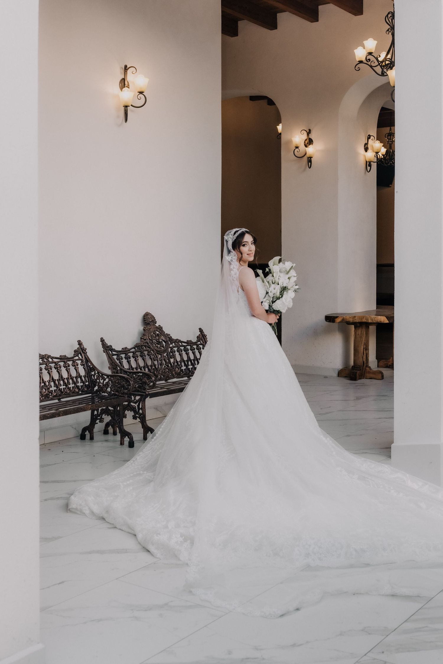 Bride in elegant white ballgown with long train holds bouquet inside ornate venue with arched columns and wall sconces.