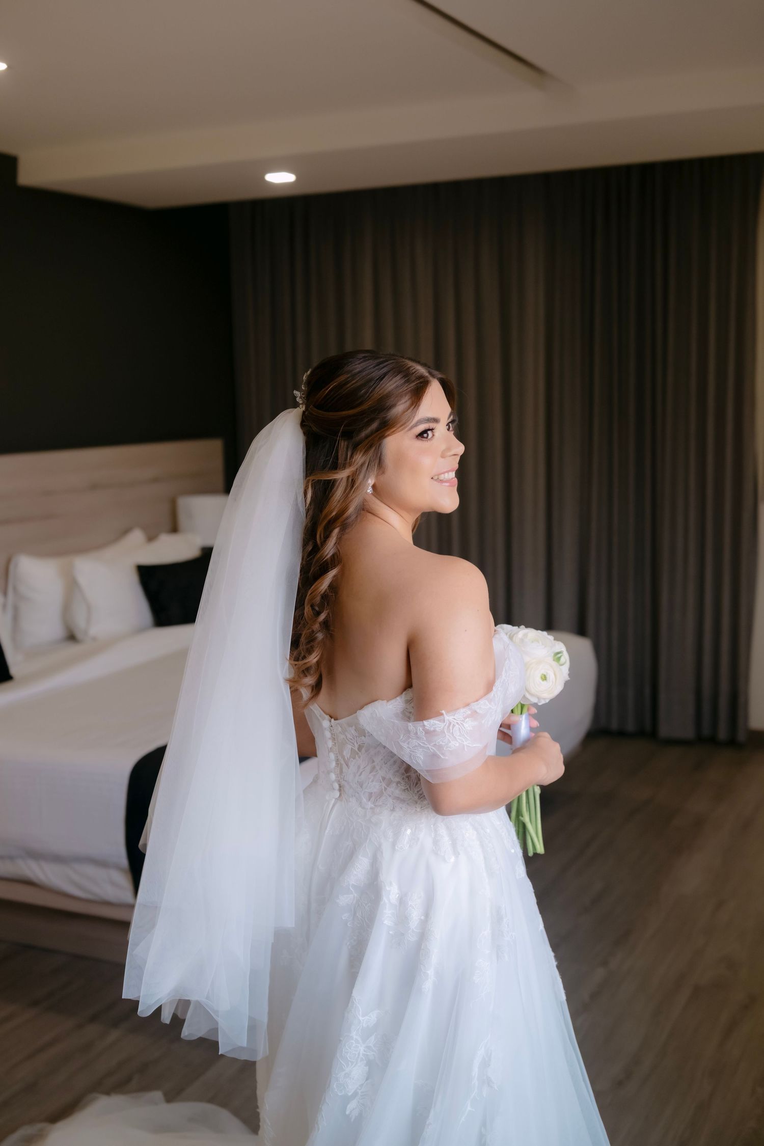 Bride in off-shoulder white gown with veil holds bouquet, smiling in elegant hotel room.