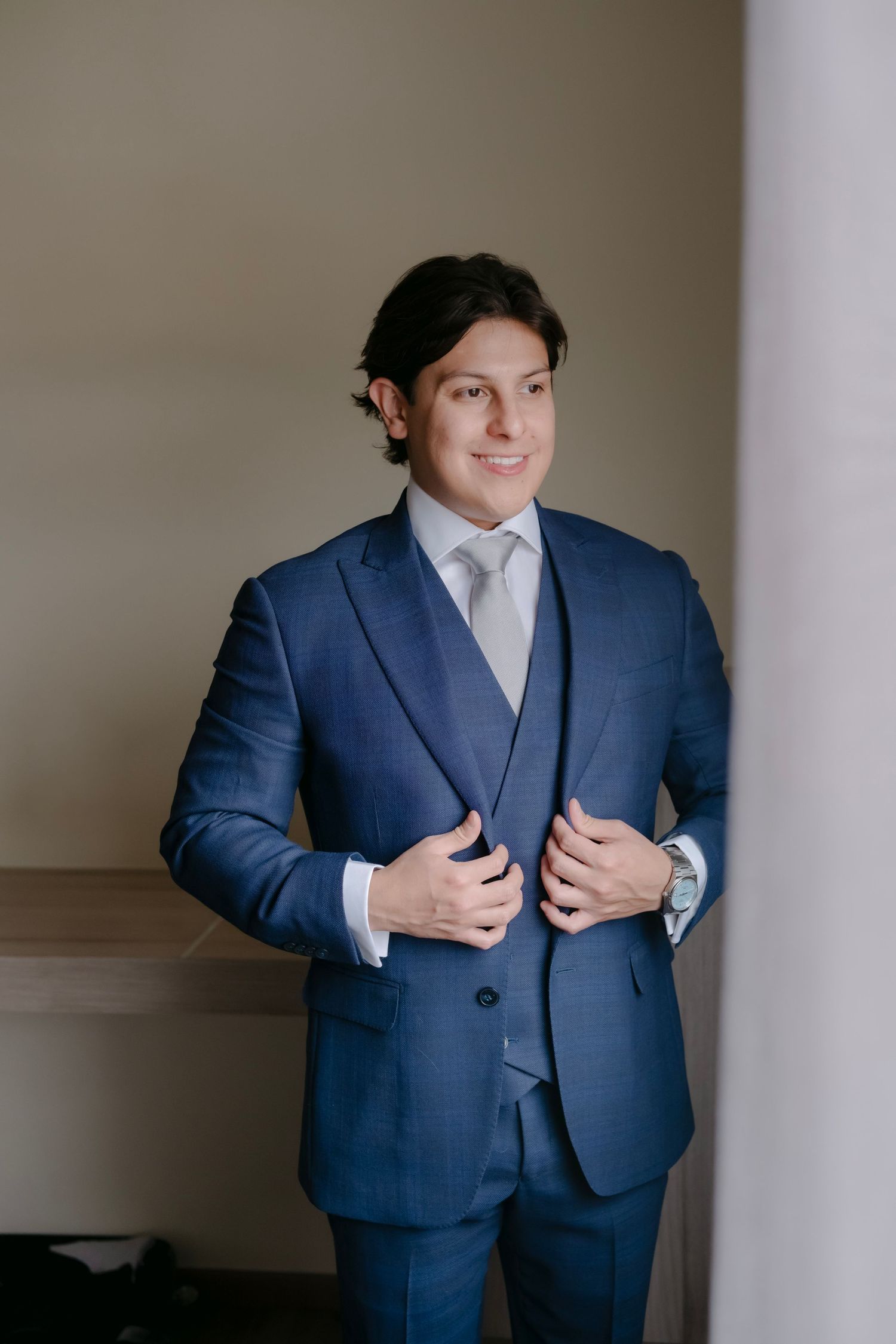 A young man in a blue three-piece suit adjusts his jacket while smiling, dressed for a formal occasion.