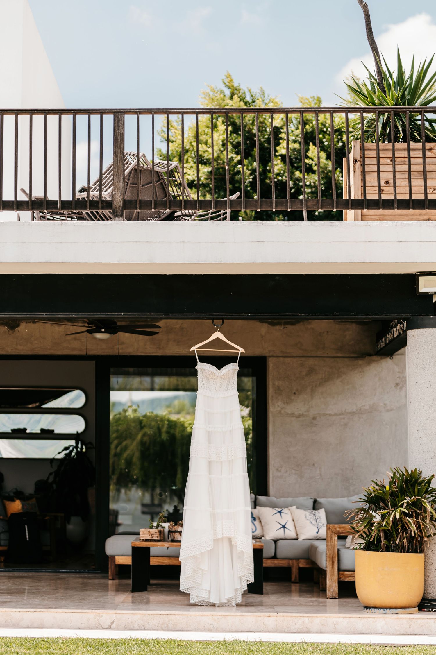 White wedding dress hanging on a hanger beneath a modern outdoor patio with lush tropical plants.