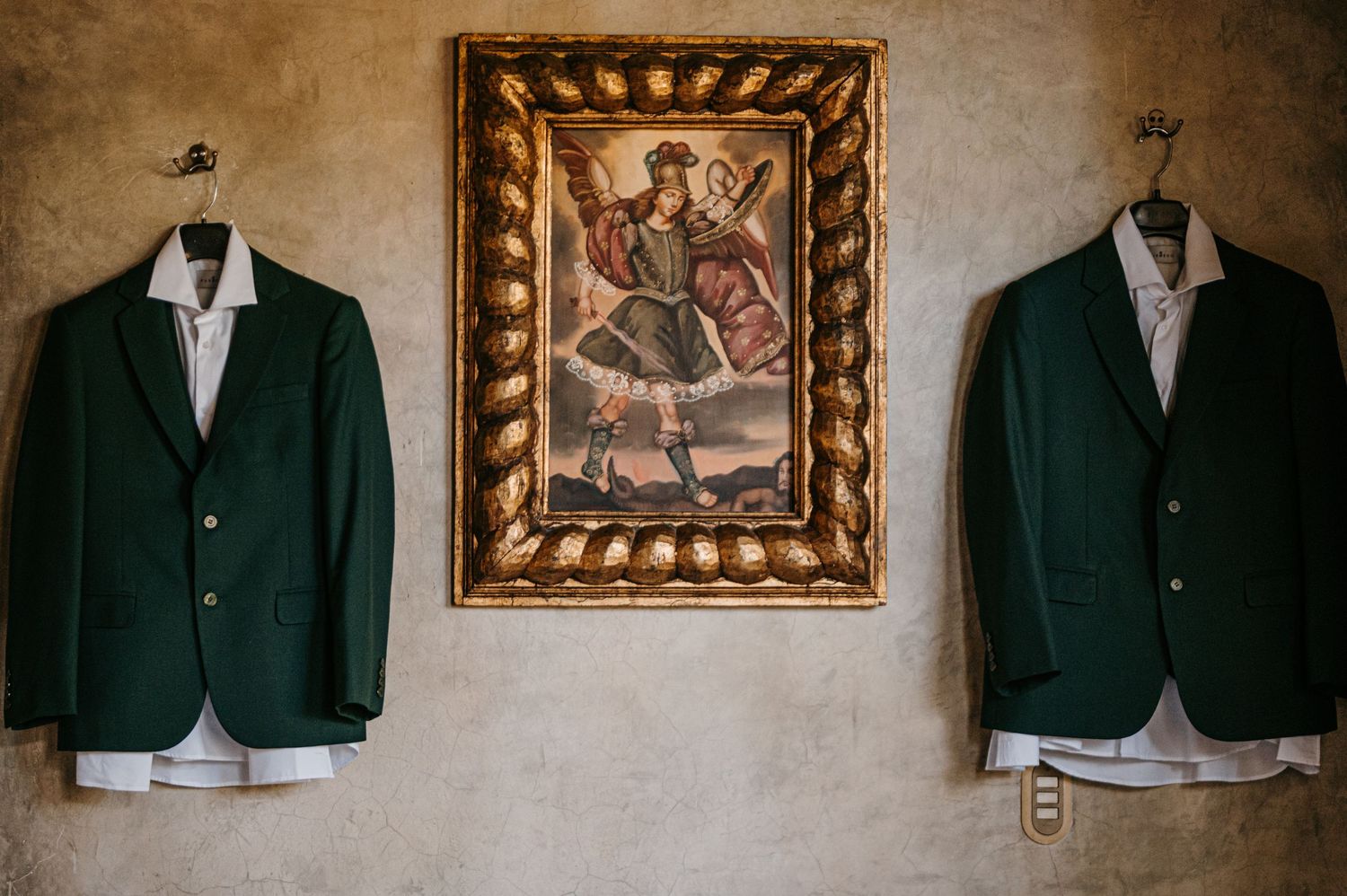 Two green blazers hanging on a wall beside a ornate gold-framed painting in an elegant vintage interior.