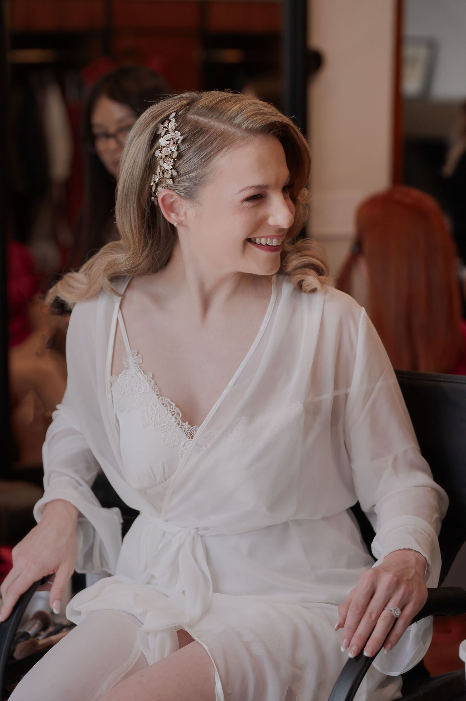 Smiling bride in white robe wearing floral hair accessory during wedding preparation.