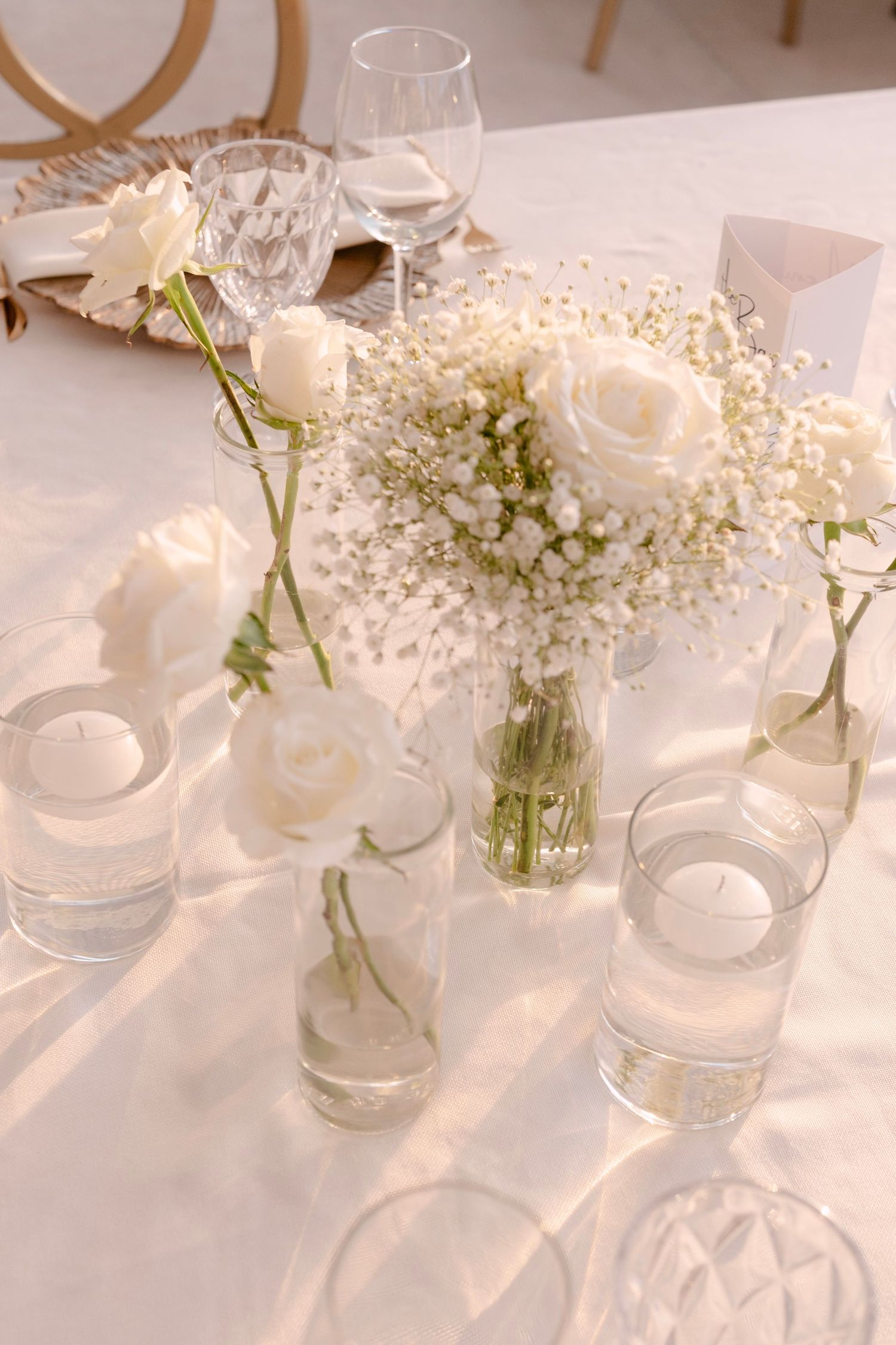 Elegant white wedding table centerpiece with roses, baby's breath in glass vases, candles, and crystal glassware.