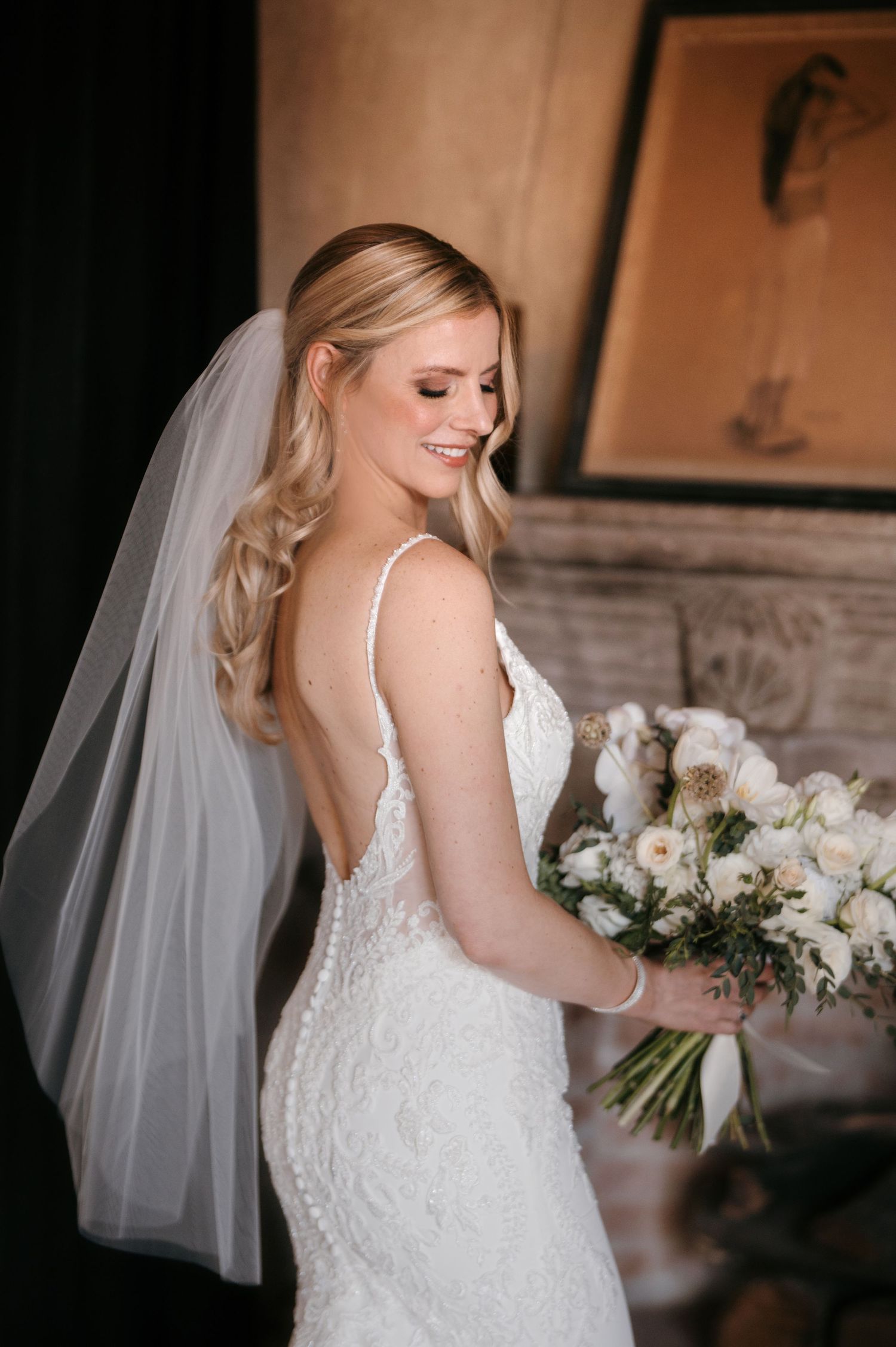 Smiling bride in backless lace gown and veil holds white bouquet, posing elegantly in a warmly lit room.