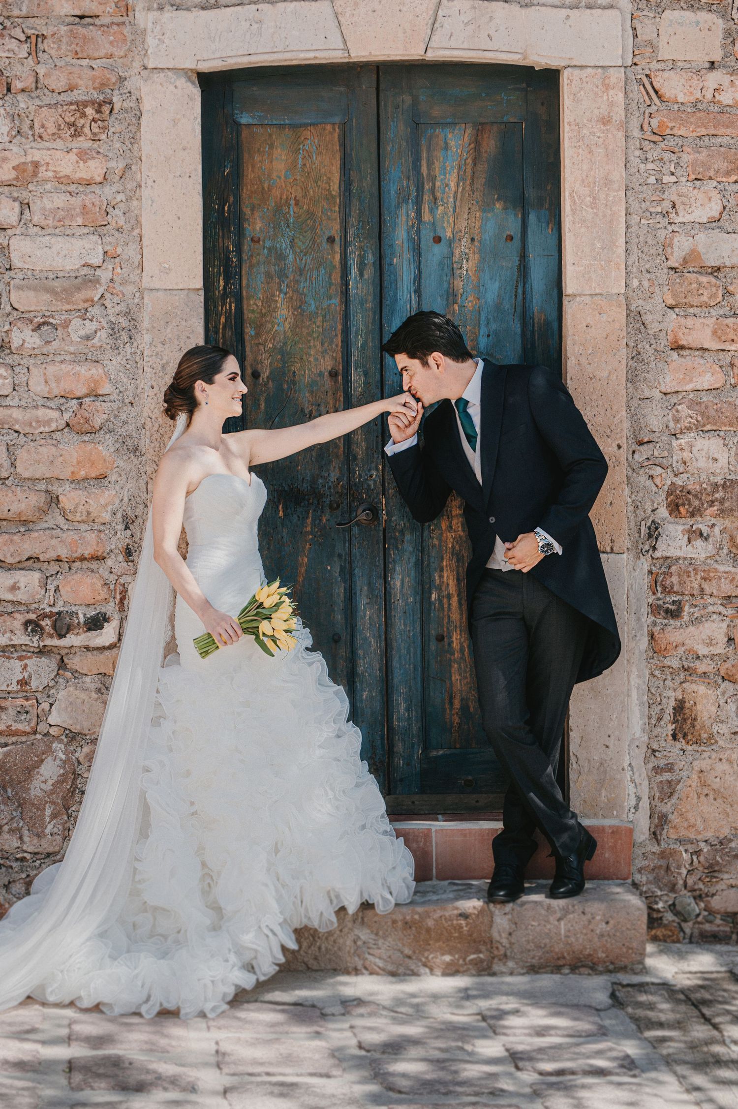 Groom kisses bride's hand in front of a rustic blue door, bride holding yellow bouquet in elegant gown.