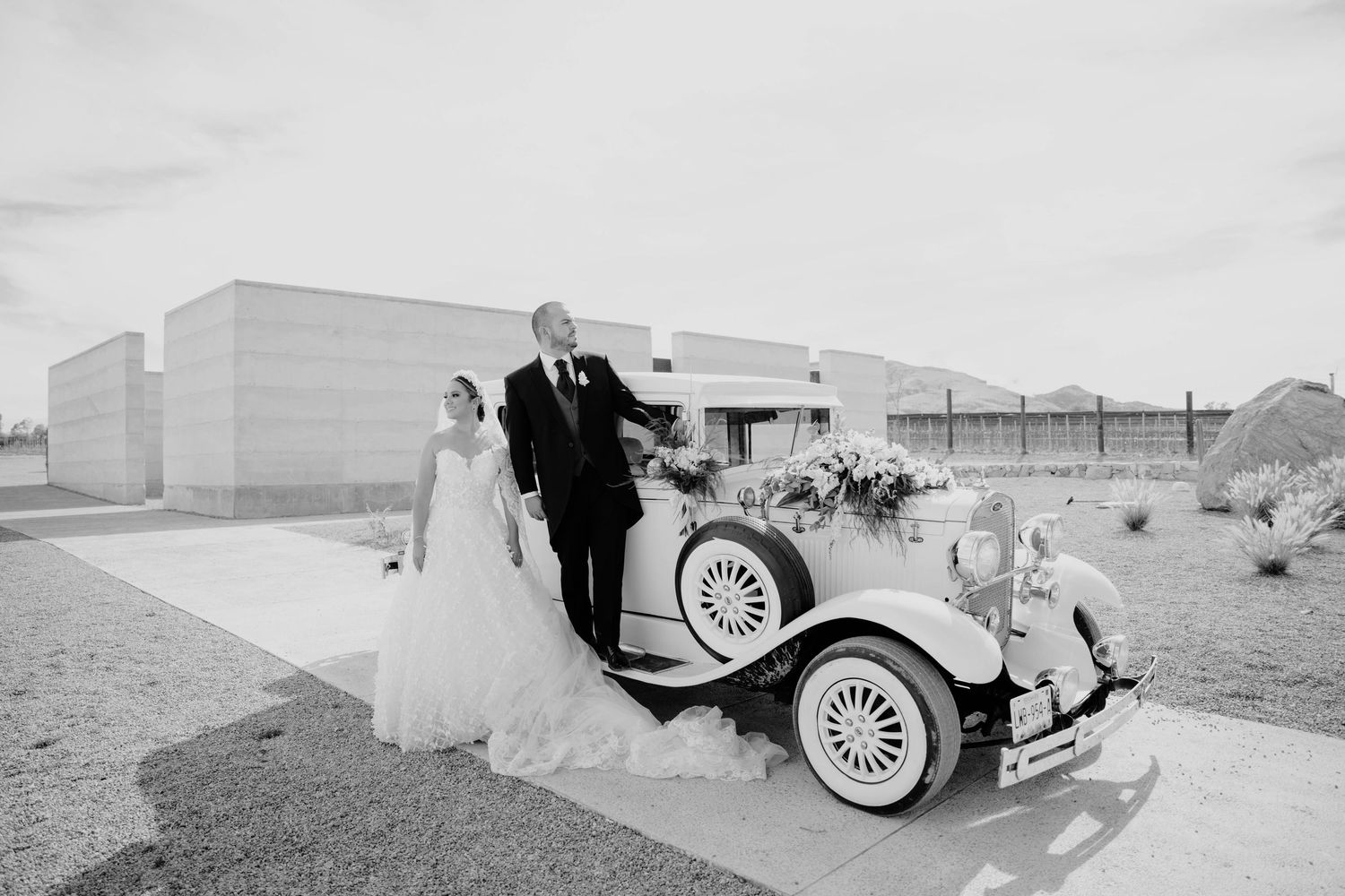 Bride and groom pose beside a vintage white classic car outside a modern wedding venue in black and white.