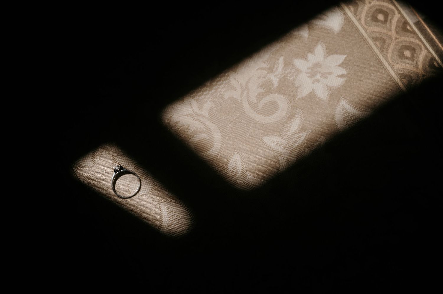 A diamond engagement ring resting in a spotlight on a dark surface beside an ornate floral patterned fabric.