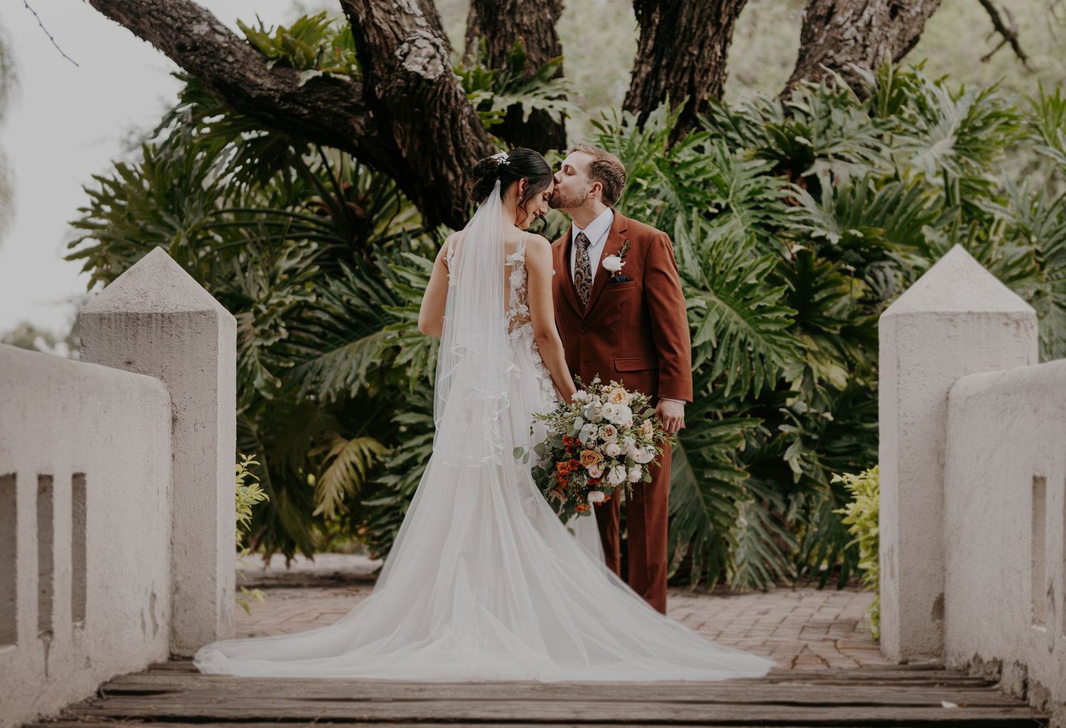 Bride and groom sharing a kiss at outdoor wedding ceremony, surrounded by lush tropical greenery.