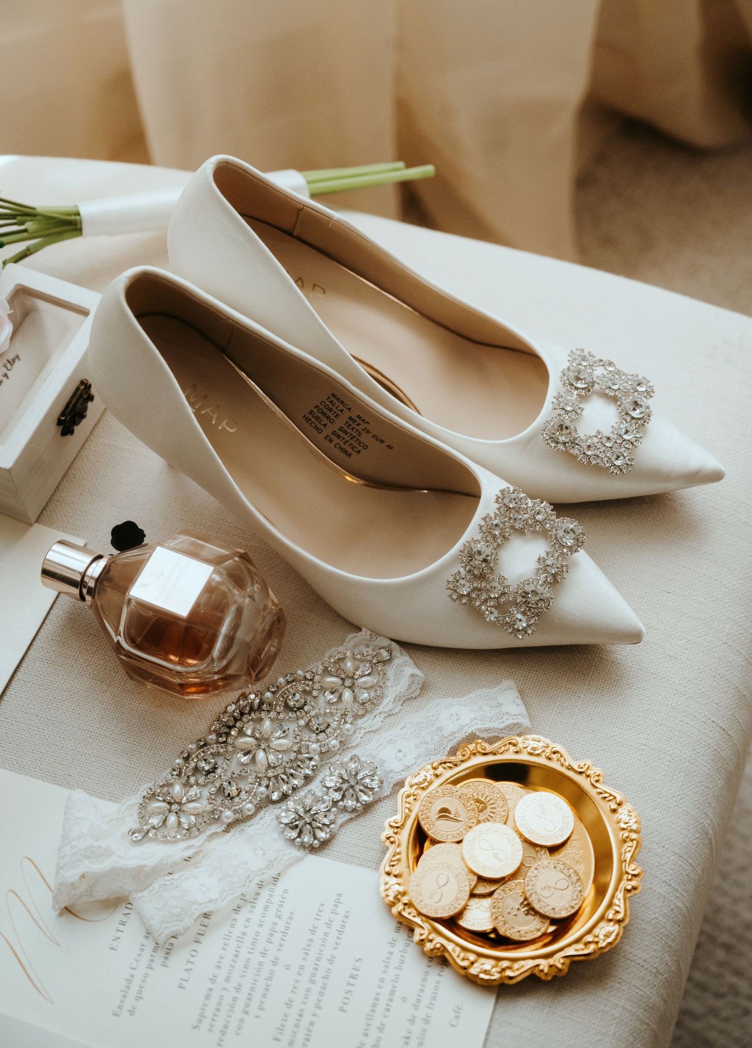 Elegant white bridal flats with crystal buckles displayed beside perfume, coins, and jewelry accessories.