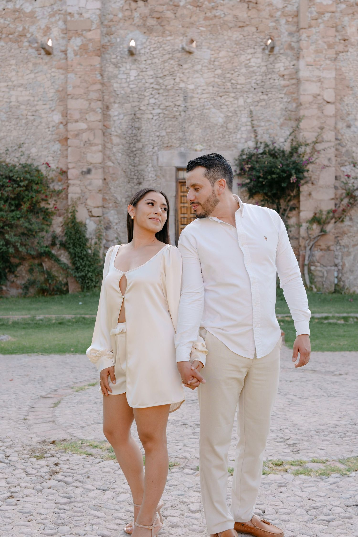 Couple in matching cream outfits holding hands and walking in front of an ancient stone wall.