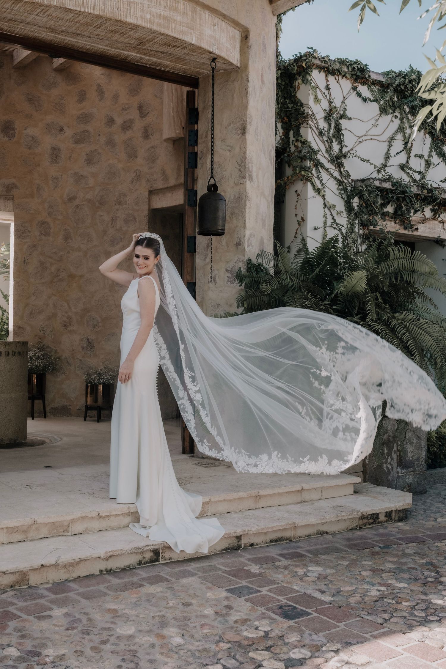 Bride in white gown with flowing lace cathedral veil billowing in wind on stone steps at rustic venue.
