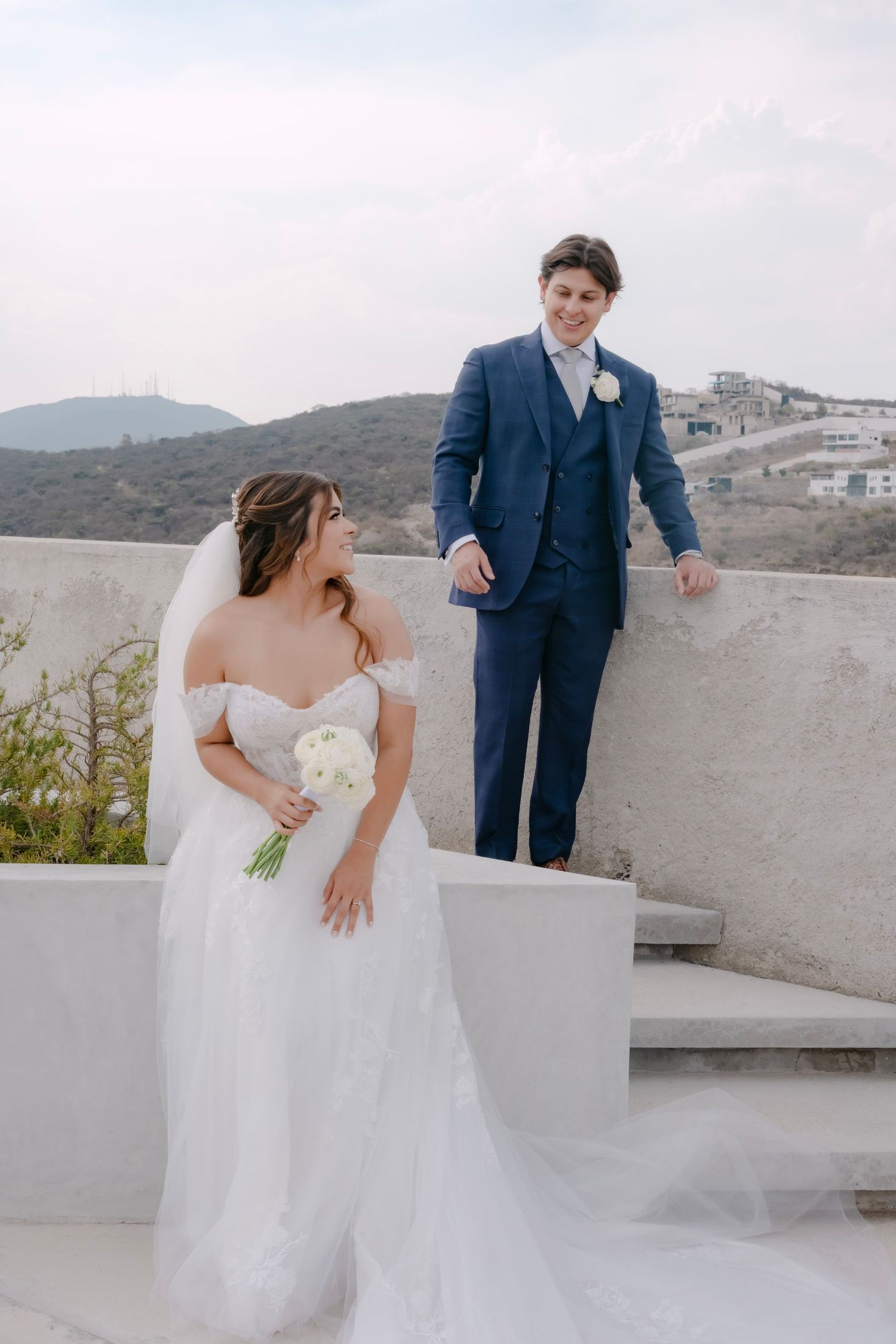 Bride in off-shoulder white gown holds bouquet as groom in navy suit smiles on outdoor terrace with hills behind.