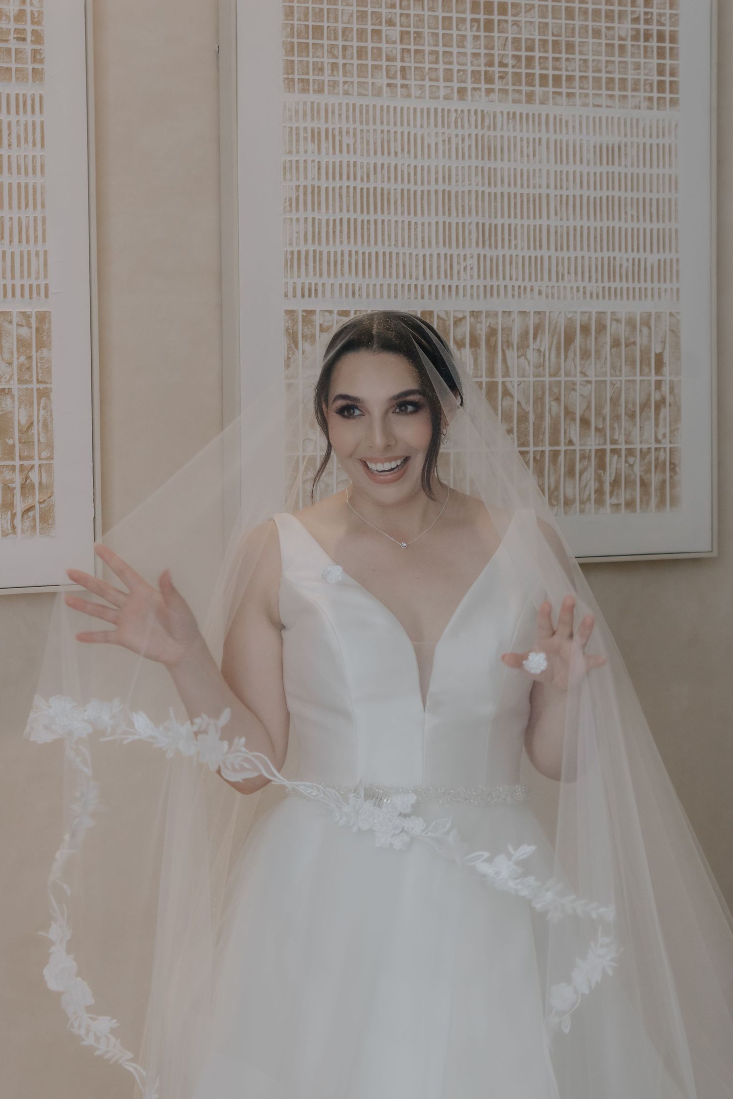 Smiling bride in white ball gown and floral-trimmed veil poses gracefully in elegant cream-toned bridal suite.
