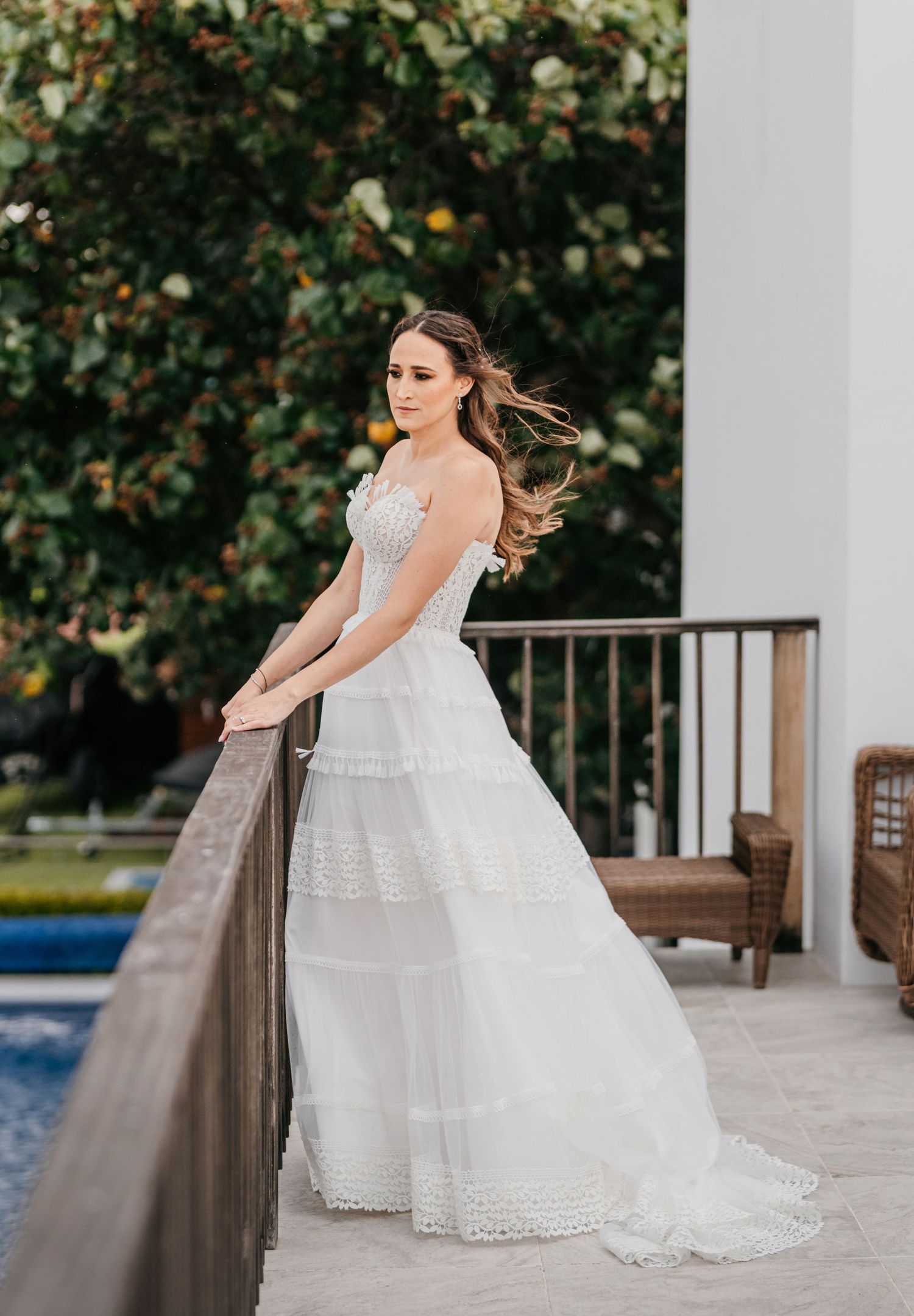 Bride in white A-line gown poses on balcony overlooking pool, hair flowing in breeze.