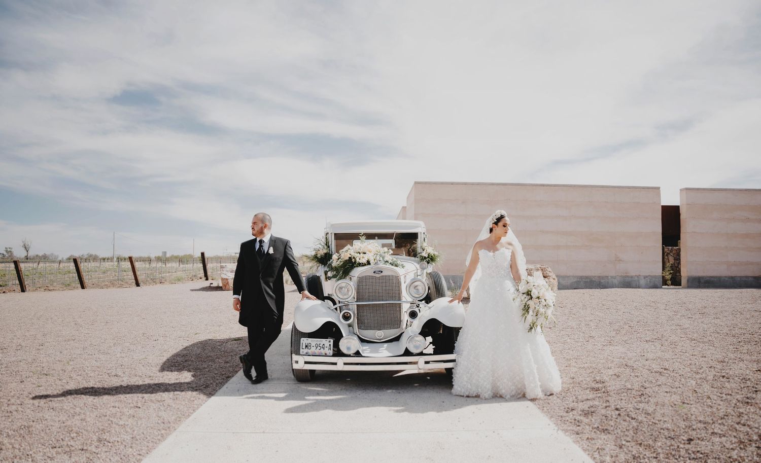Bride in white gown walks with groom beside a vintage white classic car outside a modern wedding venue.