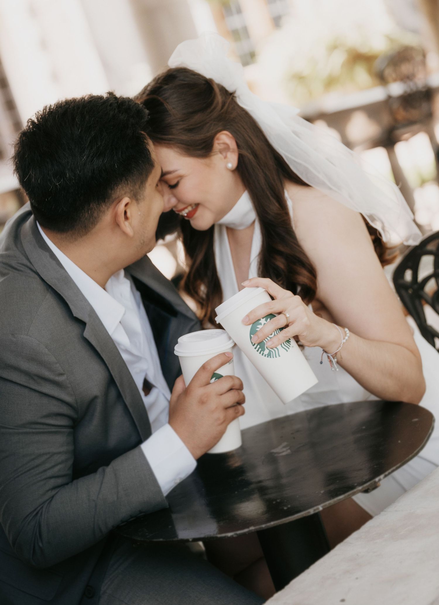 Bride and groom share a romantic moment at a cafe table, holding Starbucks cups and touching foreheads.