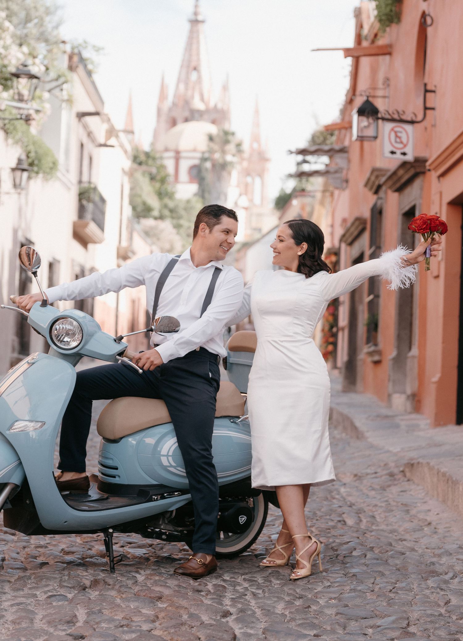 Couple posing with vintage blue Vespa scooter on cobblestone street with colorful colonial buildings backdrop.