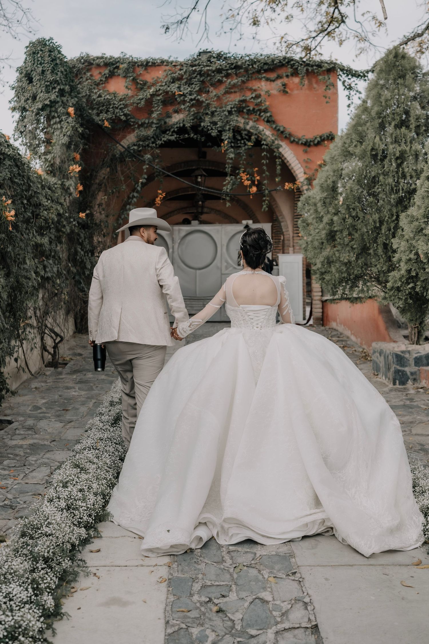 Bride in a voluminous white gown and groom in a tan suit hold hands walking toward a vine-covered arch.