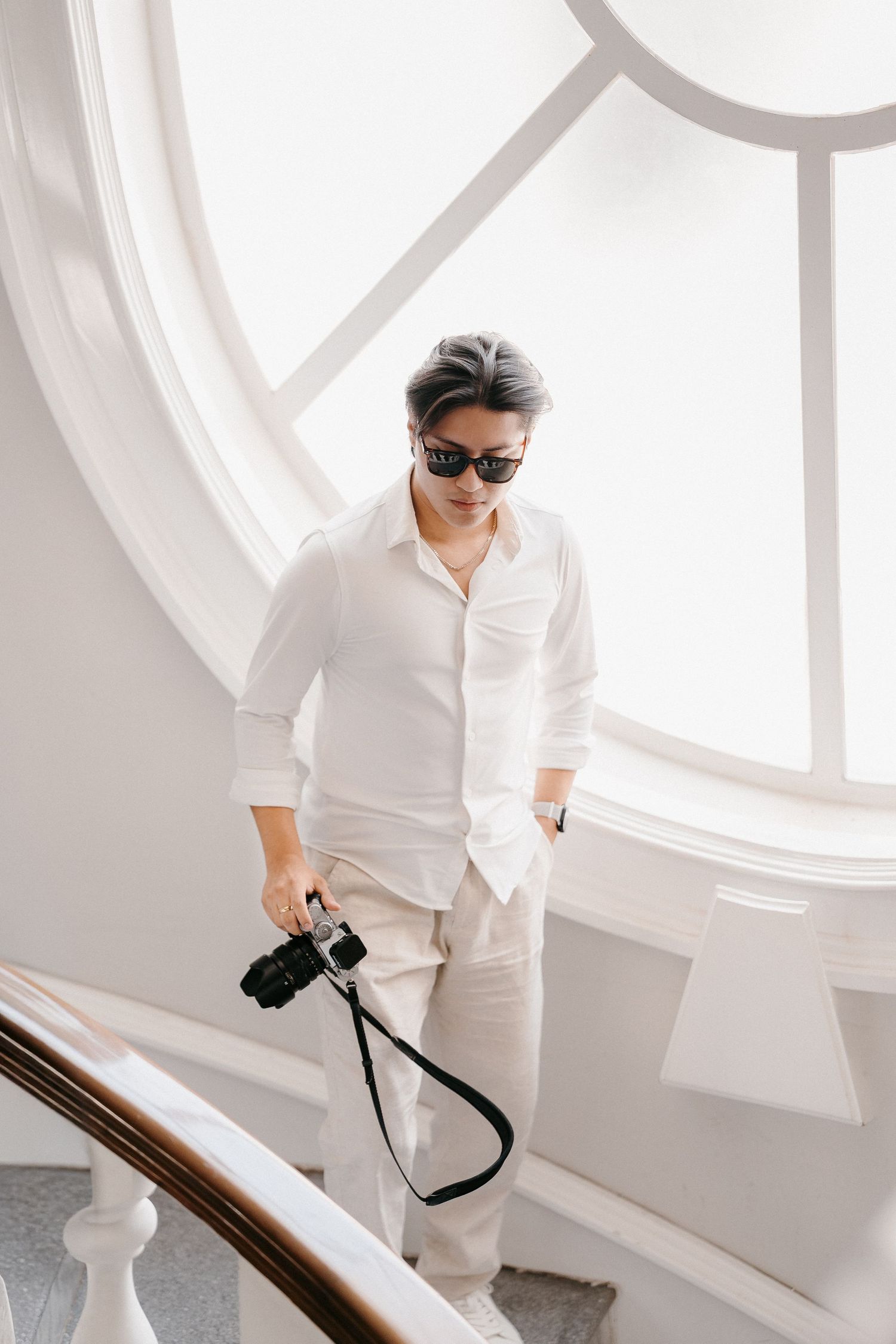 Stylish photographer in white shirt and sunglasses descending elegant staircase while holding a camera in bright interior.