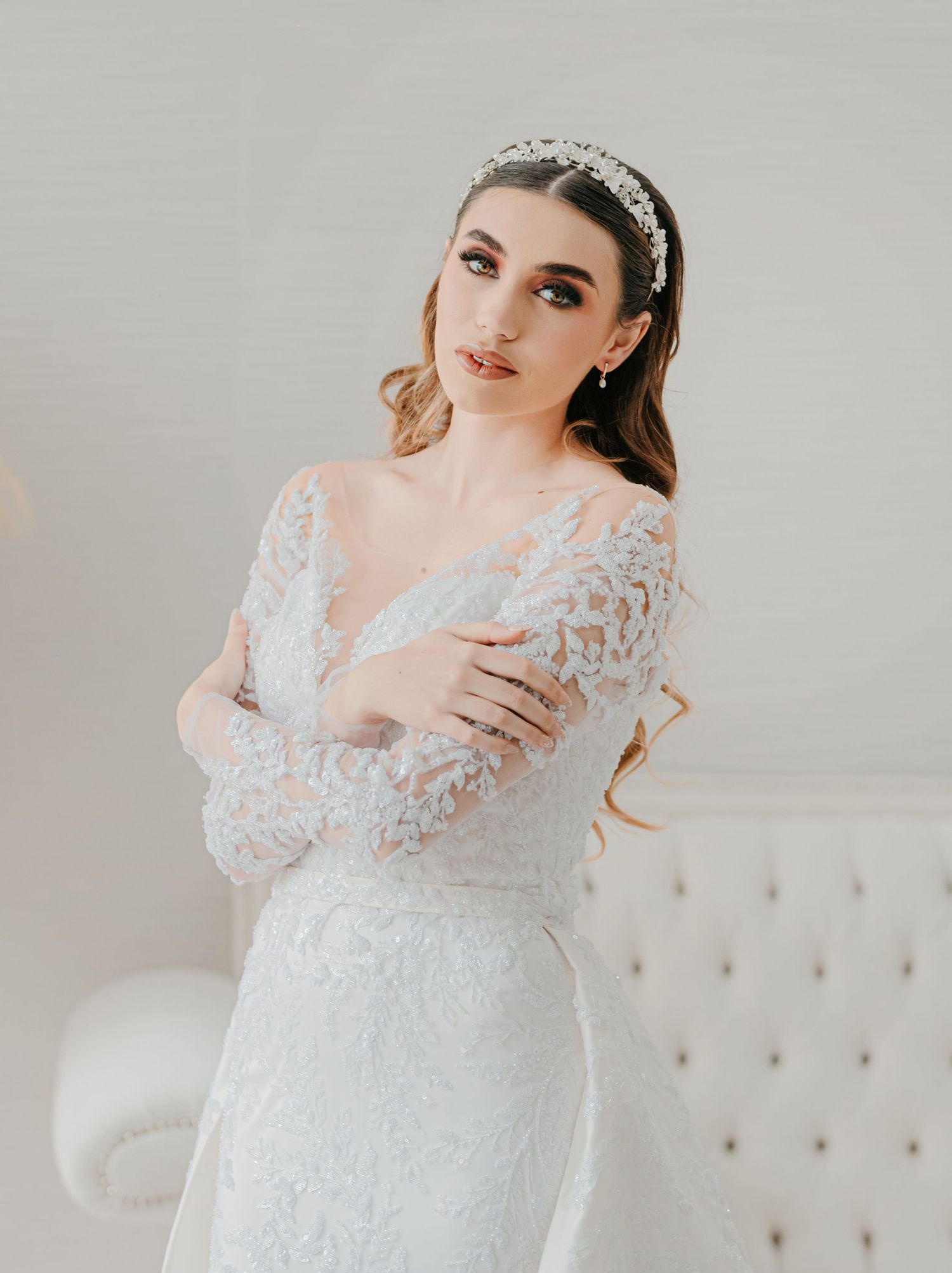 Elegant bride in long-sleeve lace wedding gown wearing crystal headband poses with arms crossed indoors.
