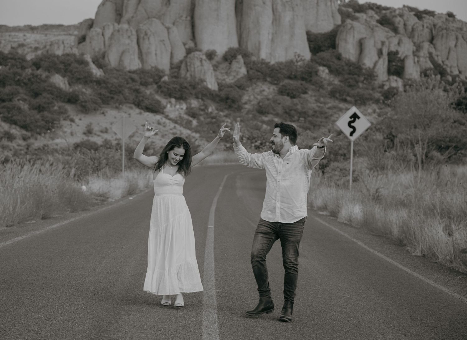 Black and white photo of a couple dancing on an empty road with rocky mountains in the background.