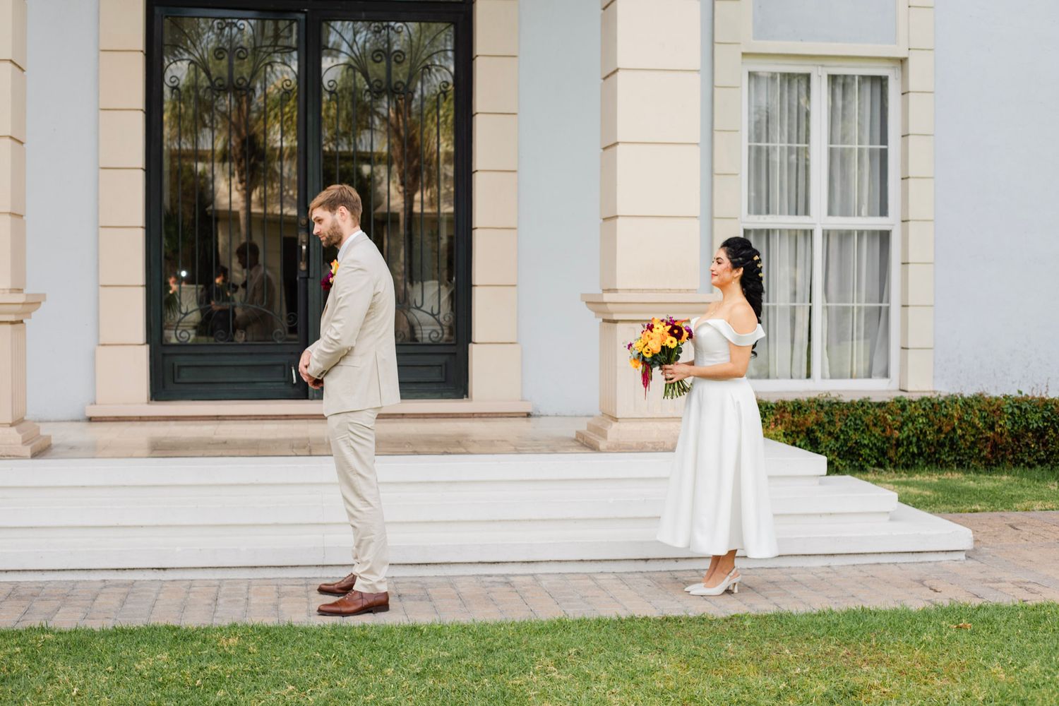 Bride holding colorful bouquet faces groom in tan suit during outdoor first look at elegant white venue.
