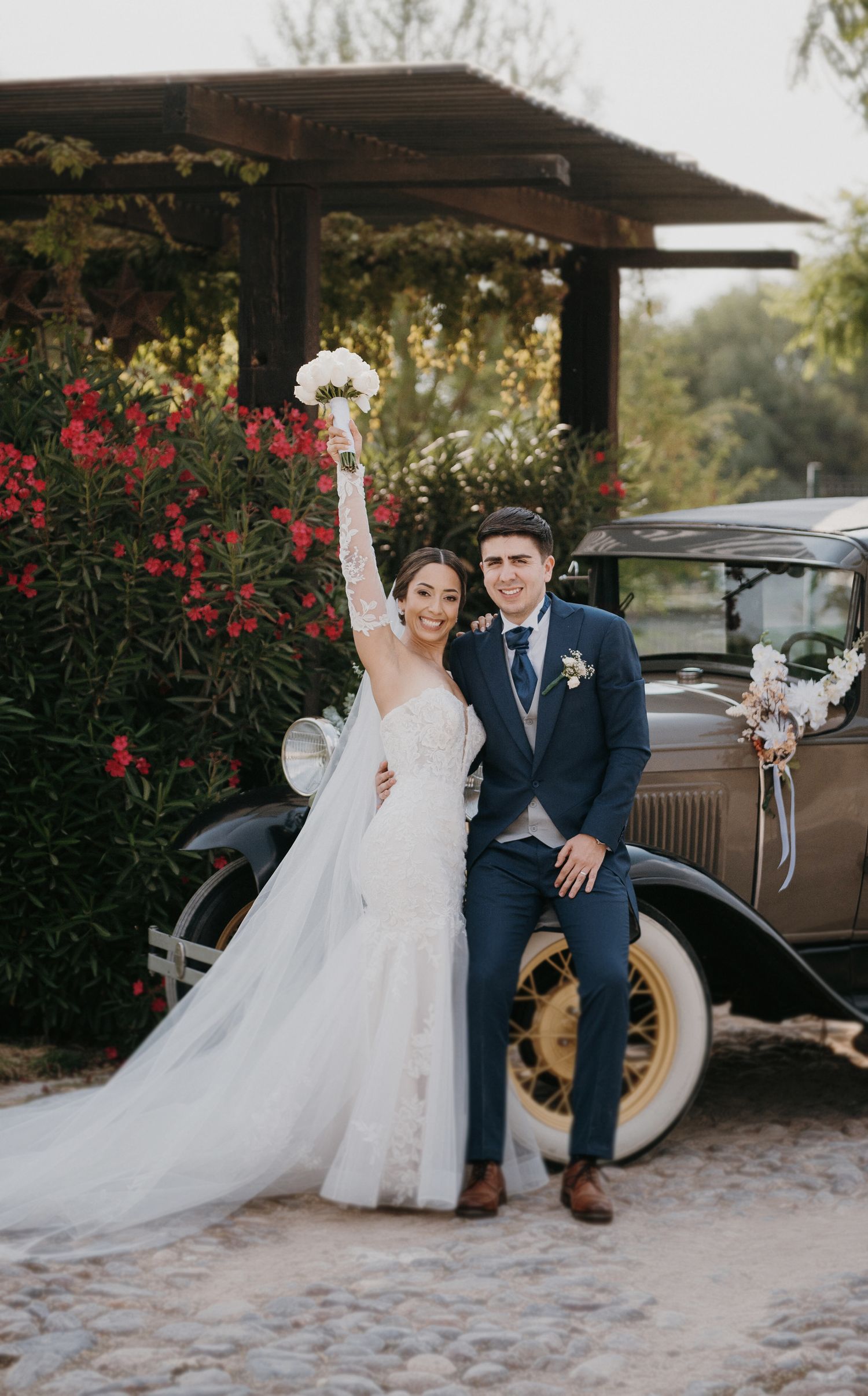 Bride and groom posing with vintage car surrounded by red flowers at wedding.
