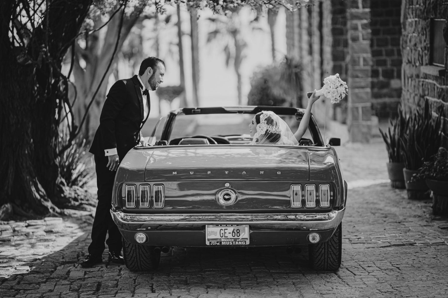 Black and white wedding photo of groom opening vintage Ford Mustang convertible door for bride holding bouquet.