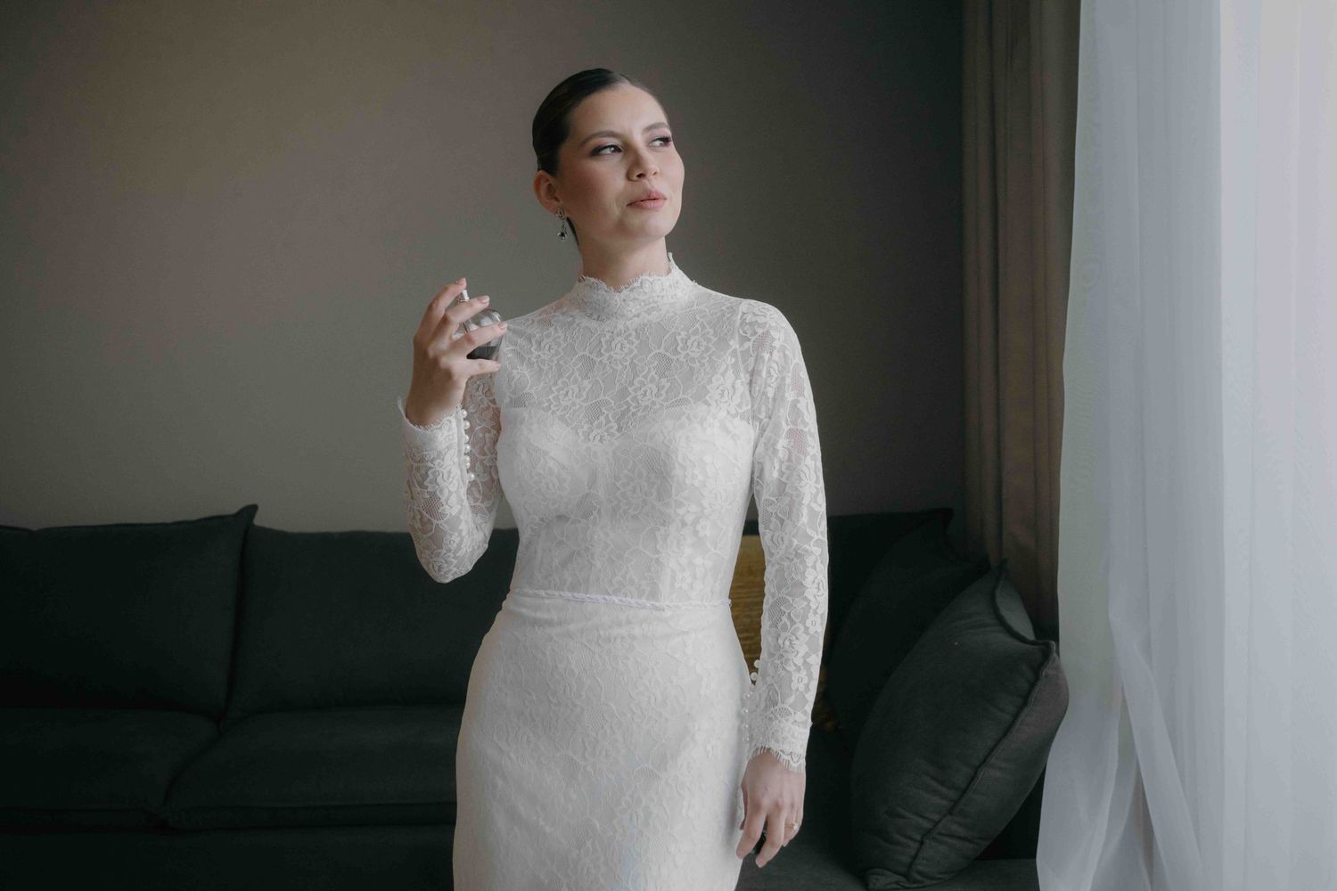 Elegant bride in a long-sleeve white lace wedding gown standing near a window in a modern room.