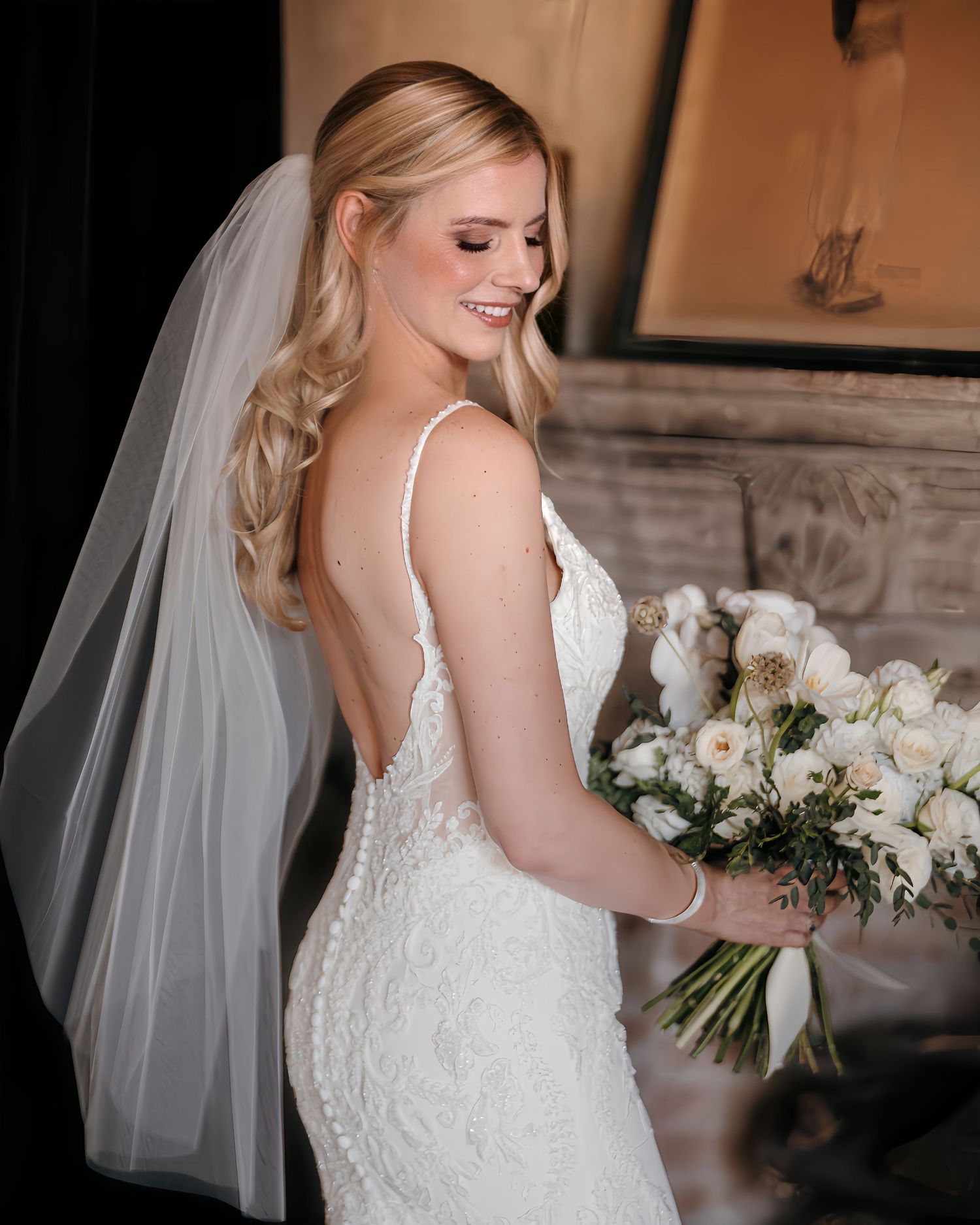Smiling bride in backless lace wedding gown with veil holding white floral bouquet.