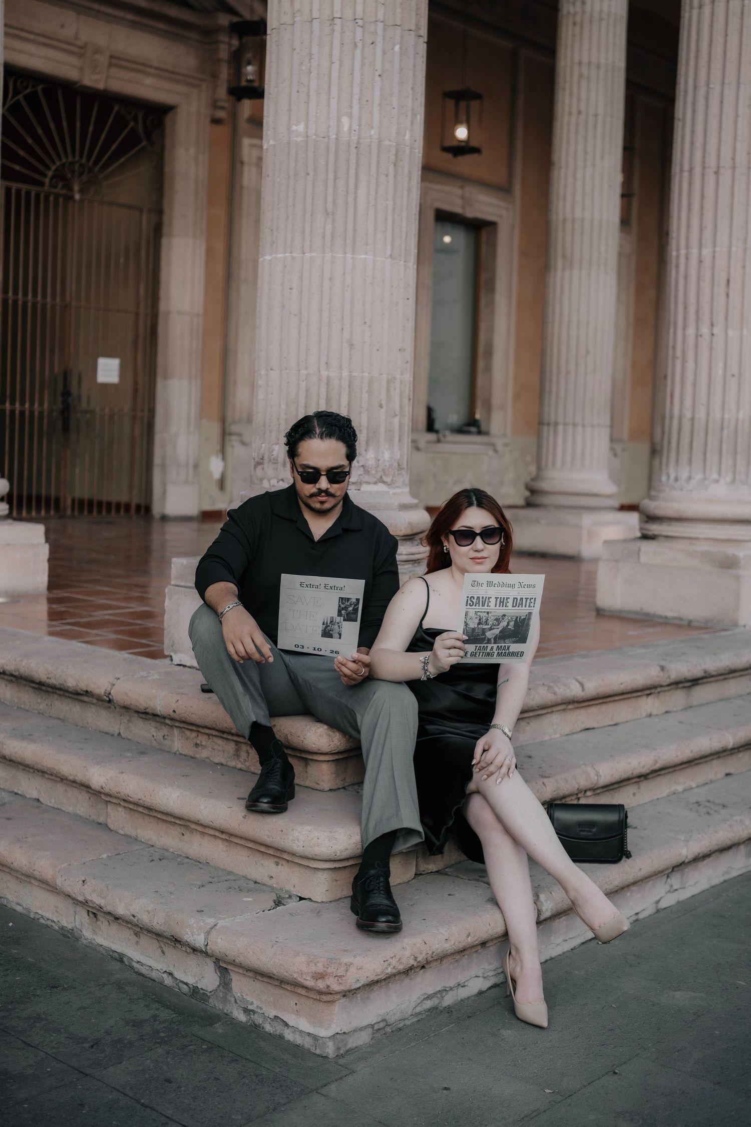 A stylish couple sits on stone steps reading newspapers, wearing sunglasses and elegant attire near classical columns.
