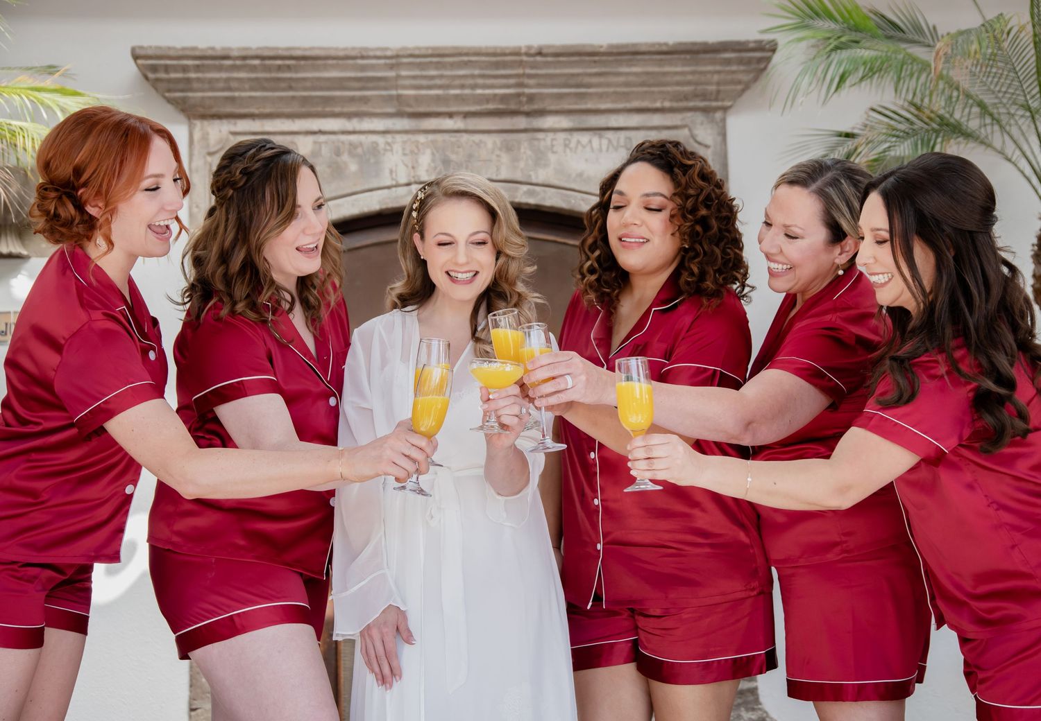 Bride in white robe toasting mimosas with six bridesmaids in matching red satin pajamas before wedding ceremony.