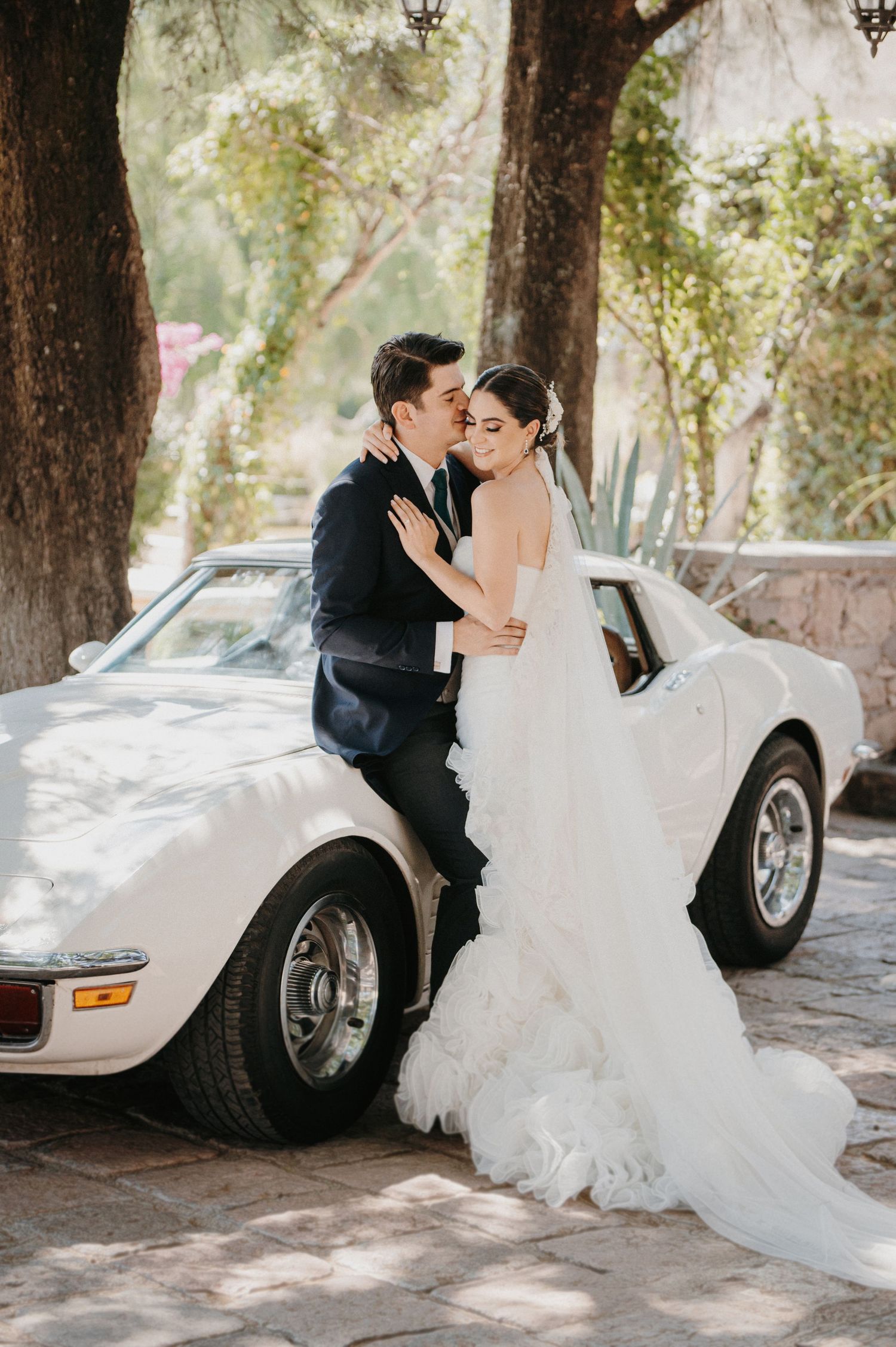 Bride and groom share a romantic kiss leaning against a classic white Corvette surrounded by lush trees.