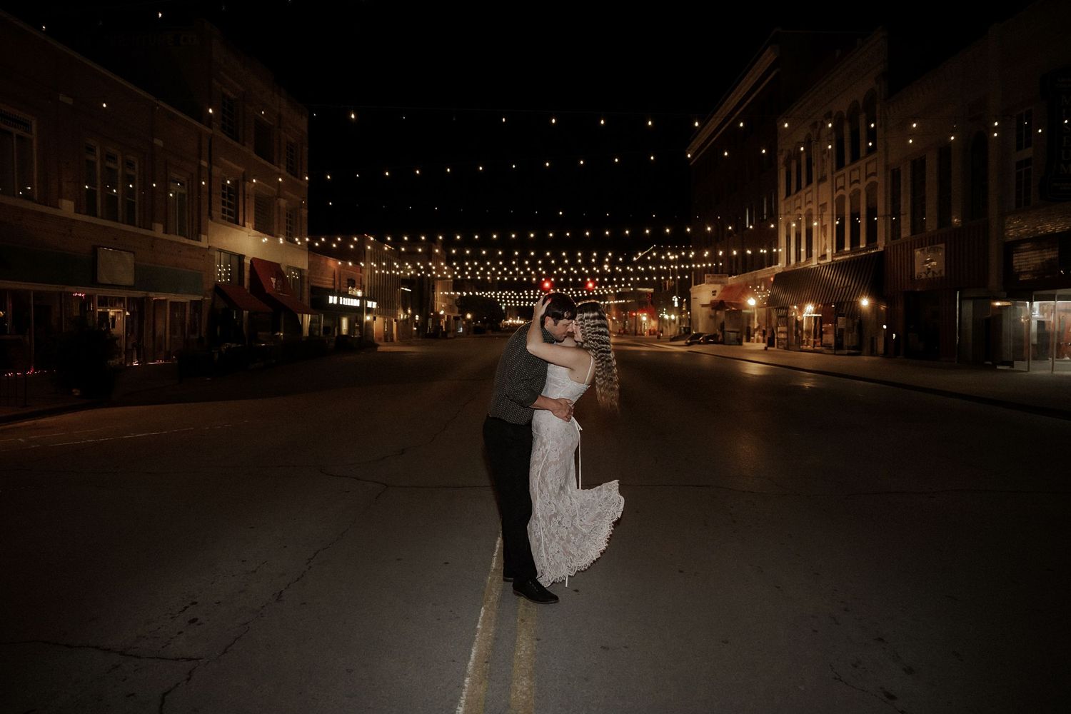 Bride and groom dance together on an empty night street lined with festive string lights overhead.