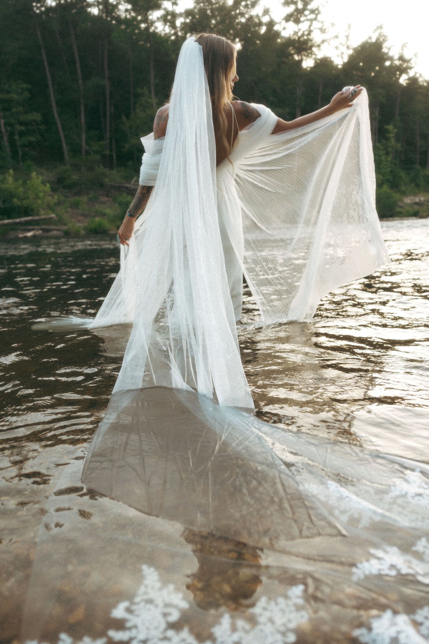 Bride standing in a shallow river holding her long lace veil wide in a forest setting.