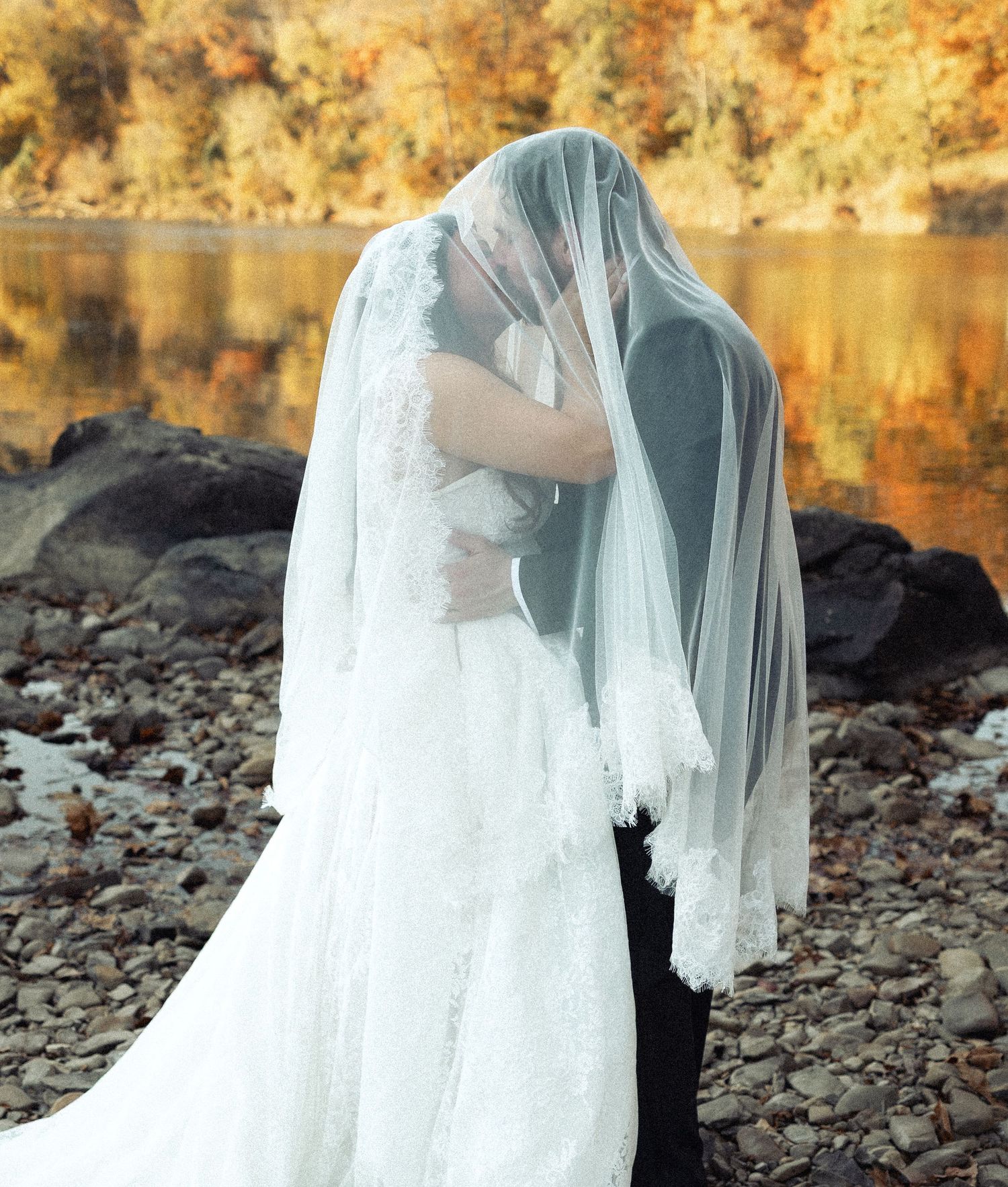 A bride and groom kiss hidden beneath a lace-edged veil on a rocky riverbank with autumn foliage.