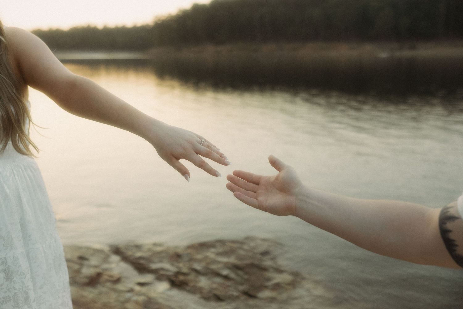 Two hands reaching toward each other near a peaceful lakeside at sunset.